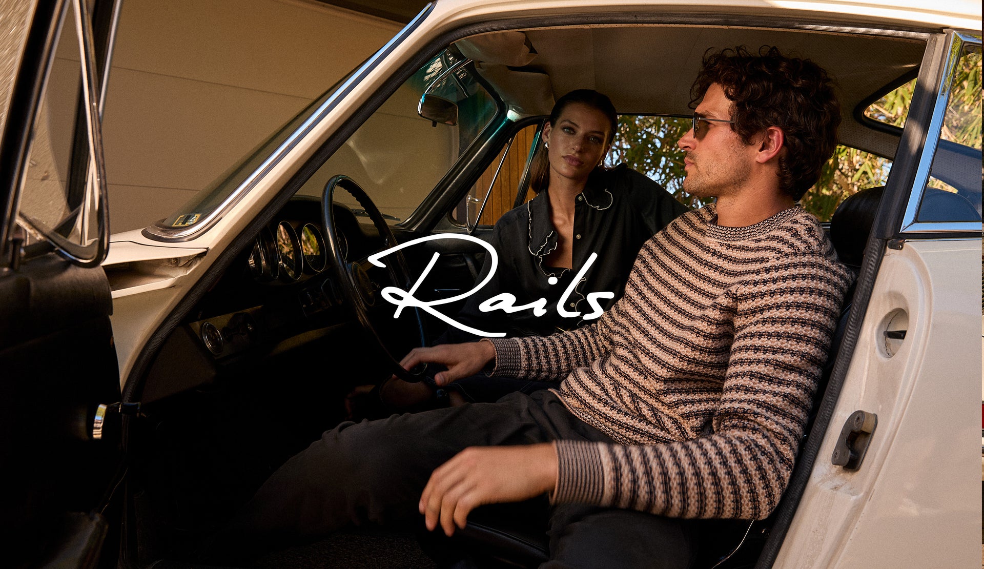 FEMALE MODEL IS WEARING FIA SHIRT (LONG SLEEVE BUTTON UP SHIRT WITH RUFFLE DETAILING AT COLLAR) IN COLOR BLACK WHITE PIPING (BLACK WITH WHITE ALONG HEM) AND DEL REY SKIRT (LONG DENIM SKIRT) IN COLOR BLACK SAND (BLACK). MALE MODEL IS WEARING CARRICK SWEATER (CREWNECK KNIT PULL OVER) IN COLOR NAVY OAT STRIPE (TAN WITH NAVY HORIZONTAL STRIPES) AND JULIAN PANT (STRAIGHT PANT WITH ELASTIC WAIST) IN COLOR BLACK