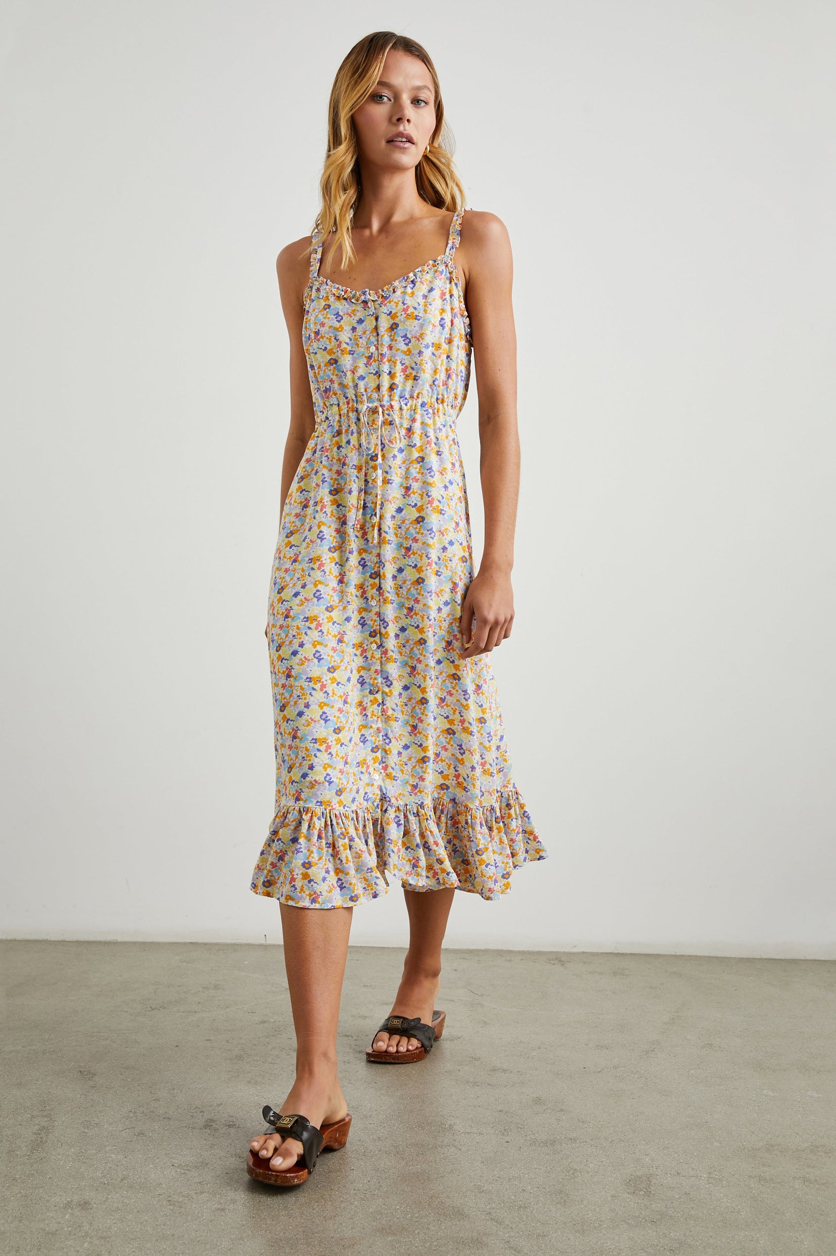 Adalyn Sleeveless Midi Dress with Thin Shoulder Straps, Buttons Down the Front, a Drawstring Waist, a Ruffled Hem and Ruffled Trim in Papaya Flowers Colorway, Purples, Blue, Yellow and Orange on White - Front Full Body View Featured on Model 