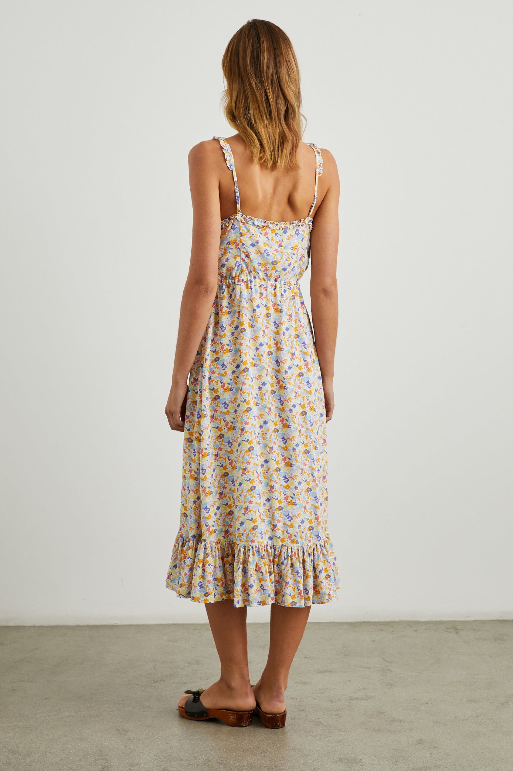 Adalyn Sleeveless Midi Dress with Thin Shoulder Straps, Buttons Down the Front, a Drawstring Waist, a Ruffled Hem and Ruffled Trim in Papaya Flowers Colorway, Purples, Blue, Yellow and Orange on White - Back Full Body View Featured on Model 