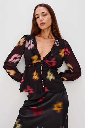 A lightweight chiffon blouse with a blurred floral print. The top features a deep V-neckline with cascading ruffles, button-front closures, and long sleeves finished with smocked bell cuffs. Front View Featured on Model.