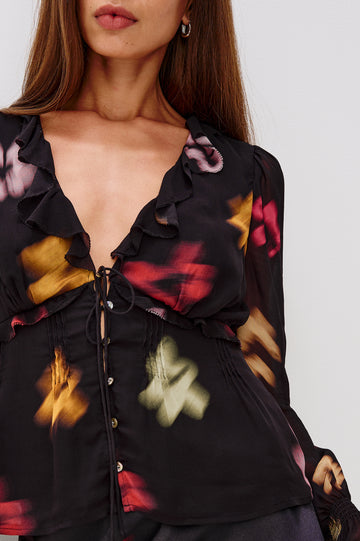 A lightweight chiffon blouse with a blurred floral print. The top features a deep V-neckline with cascading ruffles, button-front closures, and long sleeves finished with smocked bell cuffs. Close Up View Featured on Model.