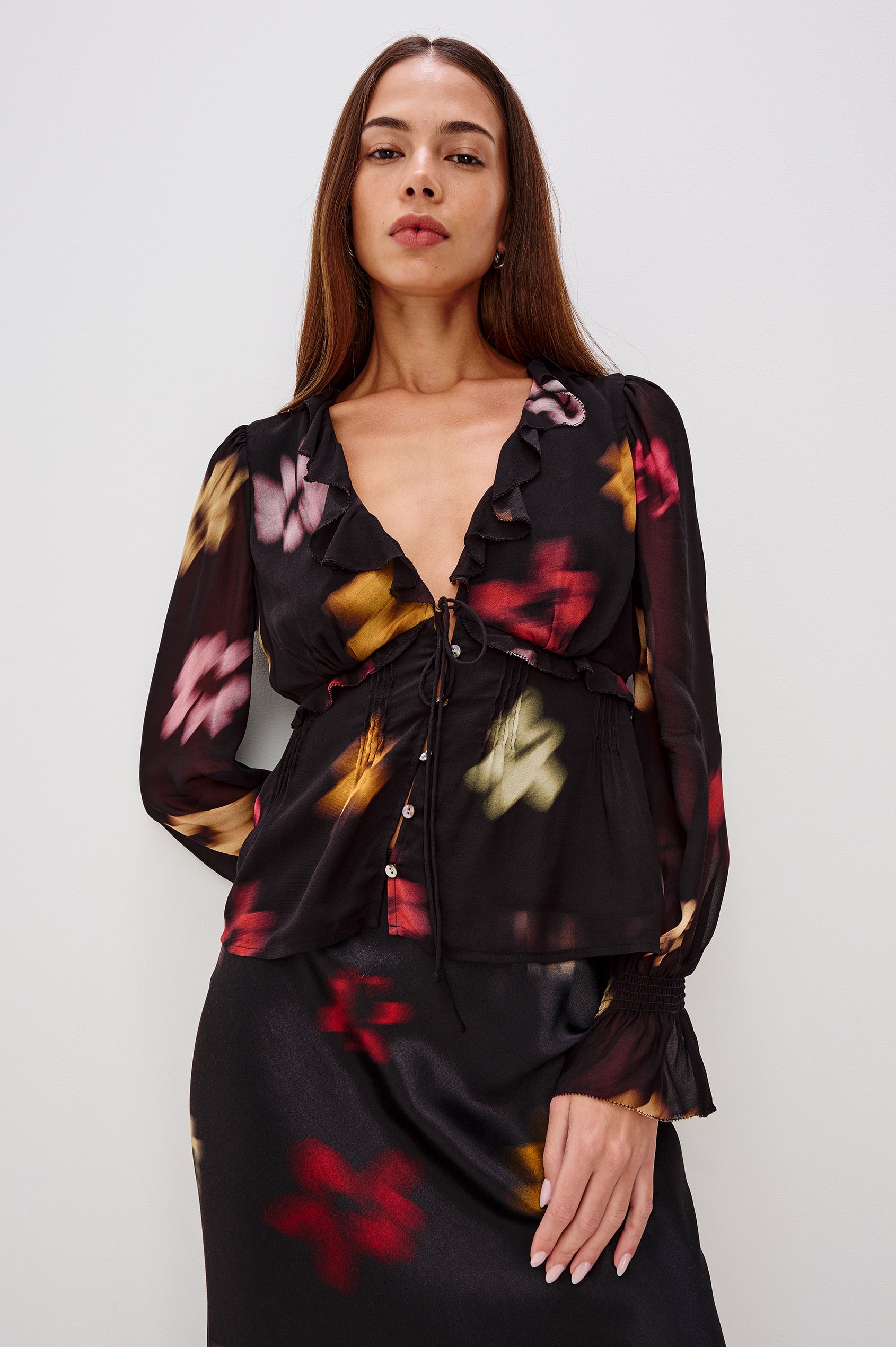 A lightweight chiffon blouse with a blurred floral print. The top features a deep V-neckline with cascading ruffles, button-front closures, and long sleeves finished with smocked bell cuffs. Front View Featured on Model.