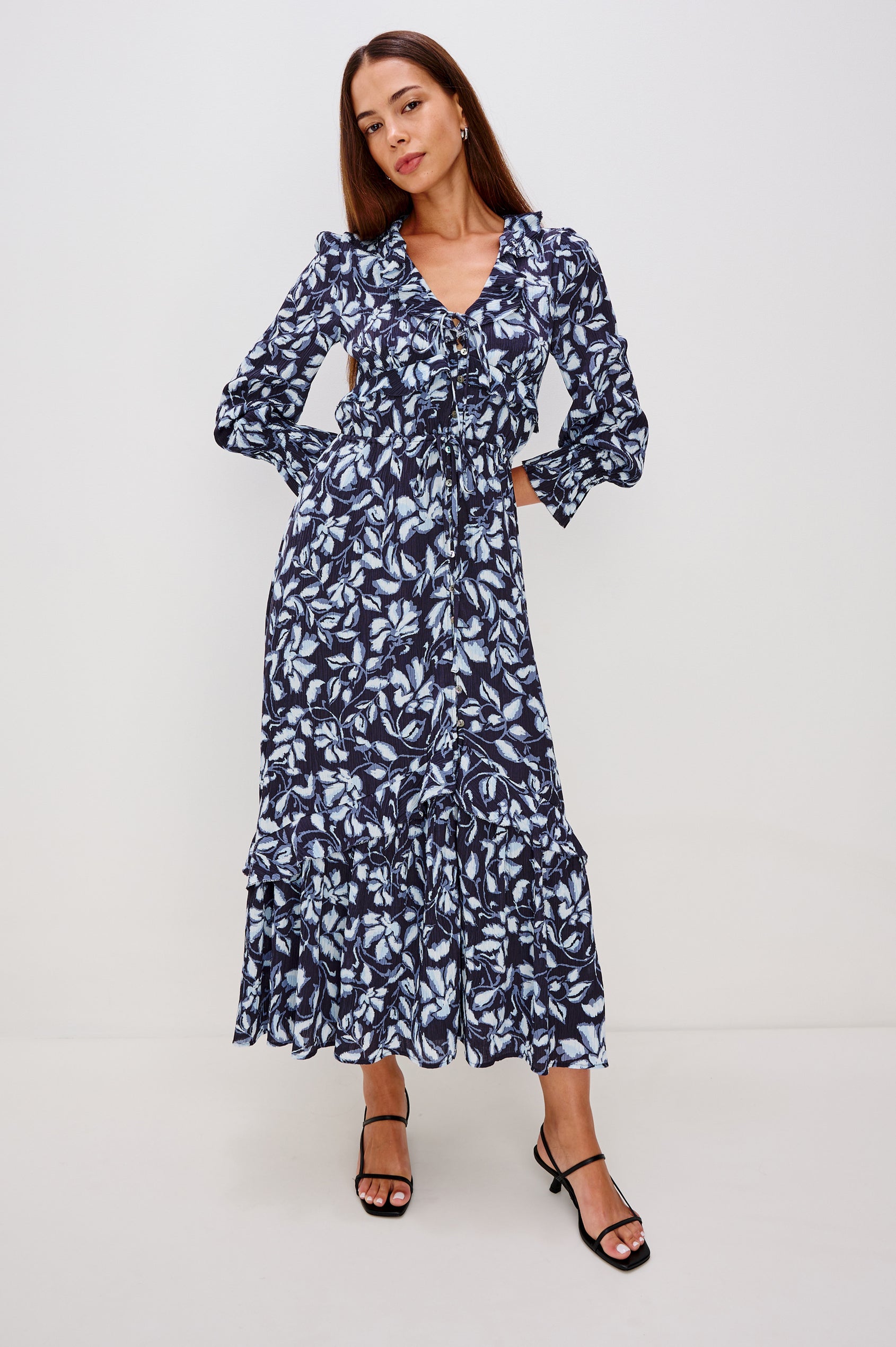 Betty is a maxi dress in pleated chiffon, it features a deep-V neckline with self ties, ruffle trim, front button-loop closures, an elastic waist with self-ties, and long sleeves with smocked cuffs in Blue Navy Jasmine Colorway.  Full body front view featured on model.