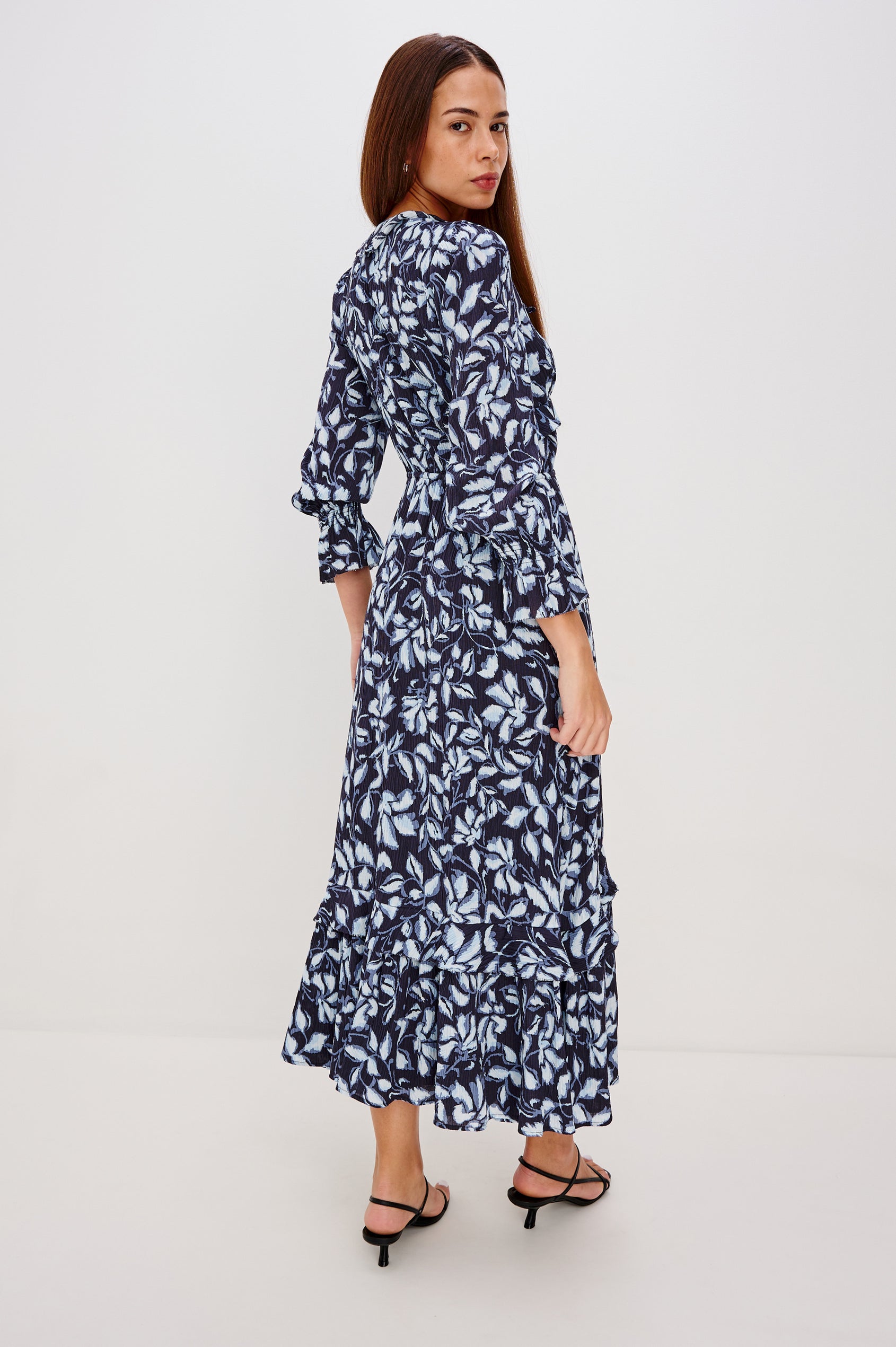 Betty is a maxi dress in pleated chiffon, it features a deep-V neckline with self ties, ruffle trim, front button-loop closures, an elastic waist with self-ties, and long sleeves with smocked cuffs in Blue Navy Jasmine Colorway.  Full body back view featured on model.