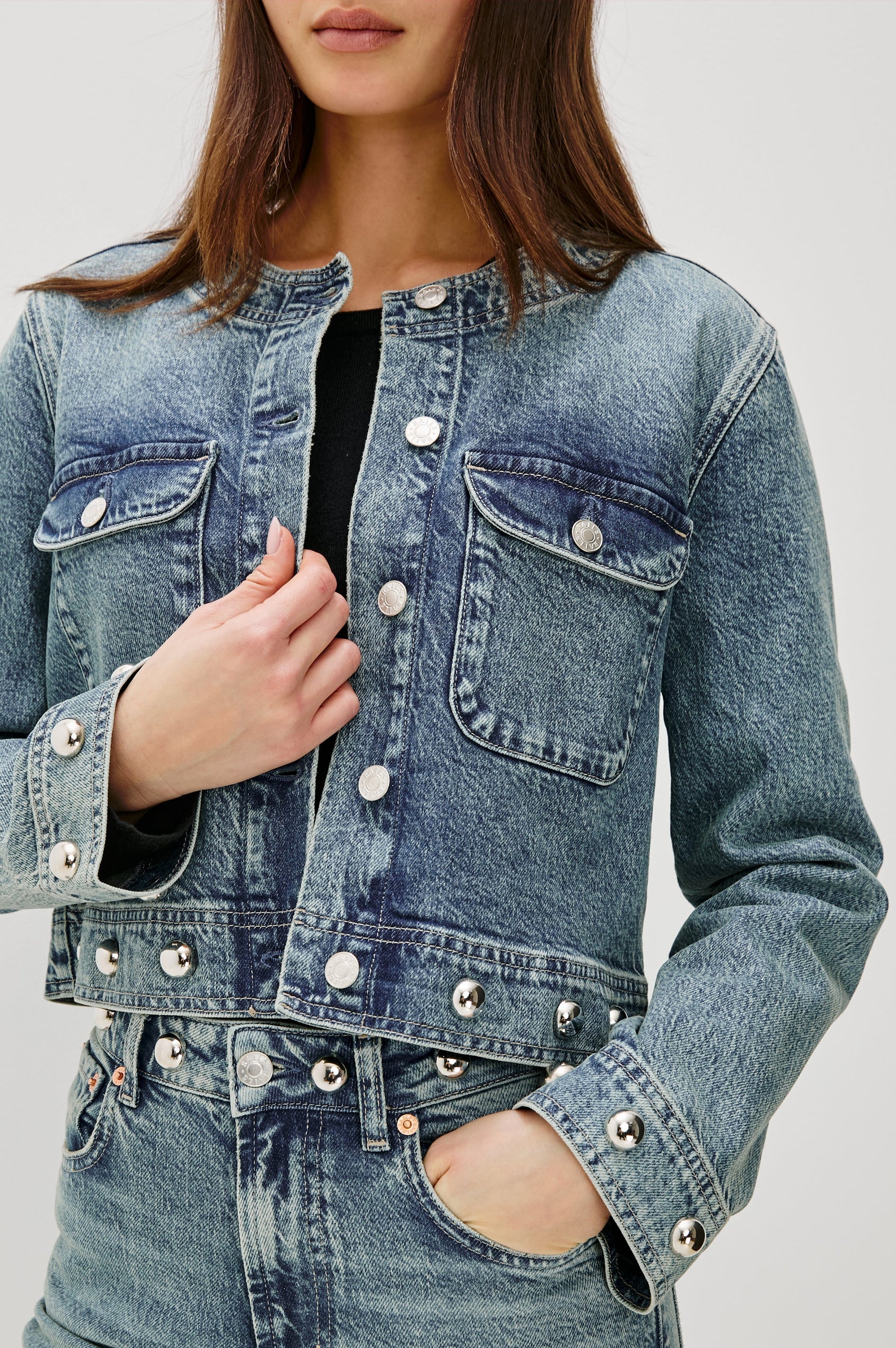 The Capistrano is a cropped denim jacket with a scoop neck, it has a button front, two chest pockets and silver studs on the sleeve cuffs and hem in Desert Sky colorway. Front detail view featured on model.