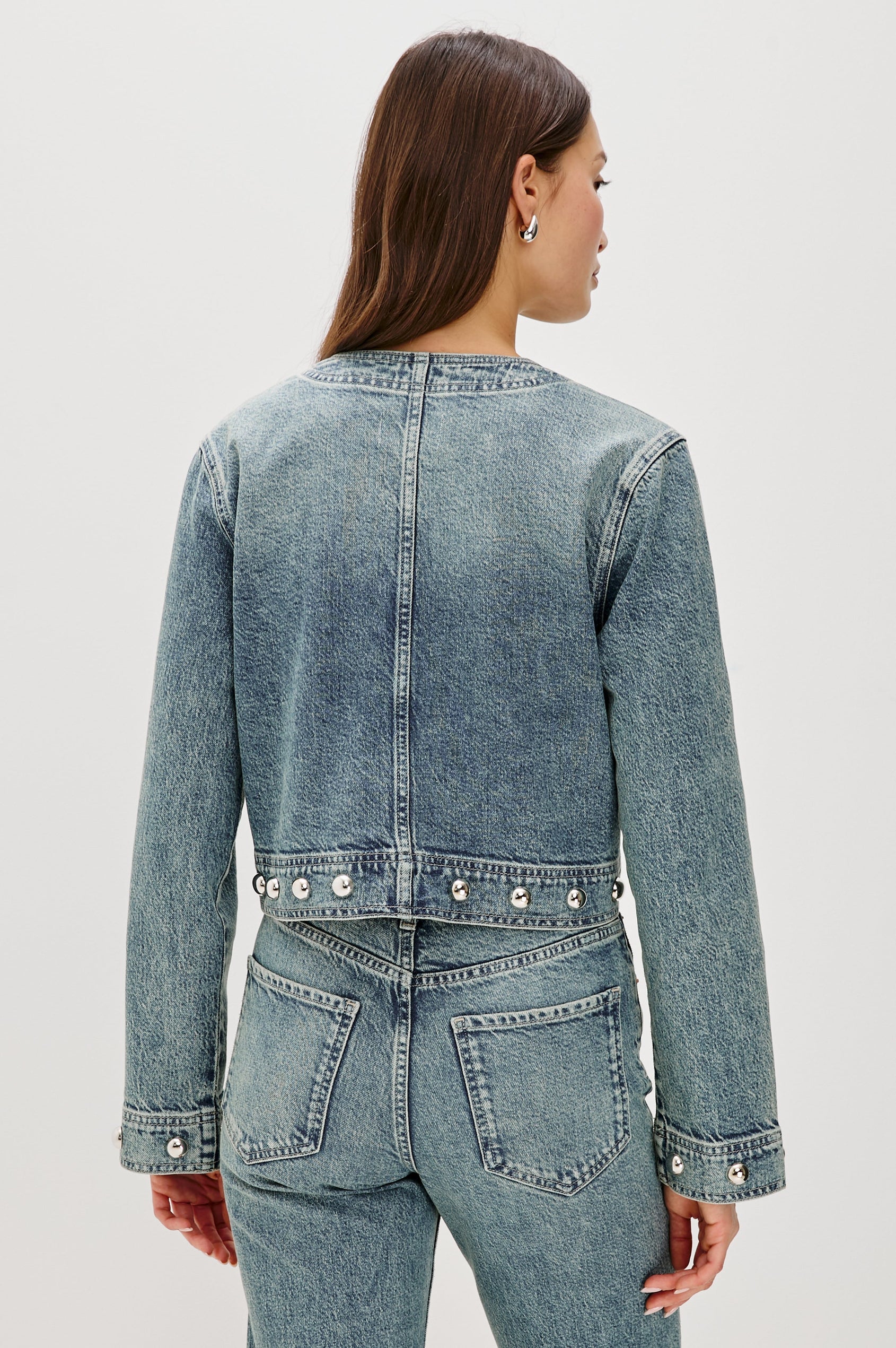 The Capistrano is a cropped denim jacket with a scoop neck, it has a button front, two chest pockets and silver studs on the sleeve cuffs and hem in Desert Sky colorway. Back view featured on model.