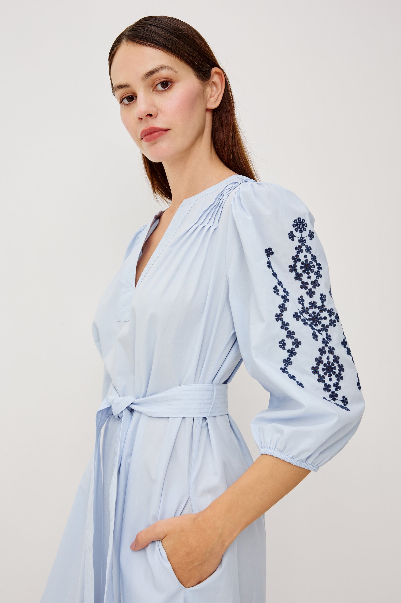 Carmel Peasant Maxi Dress in Poplin With Pleated Shoulders, V-Neckline, Three-Quarter Puff Sleeves, Adjustable Waist Ties, Side Pockets, Tiered Seam, and Contrast Embroidery at Cuffs and Hem in Blue Bird Colorway - Upper Body Side View Featured on Model