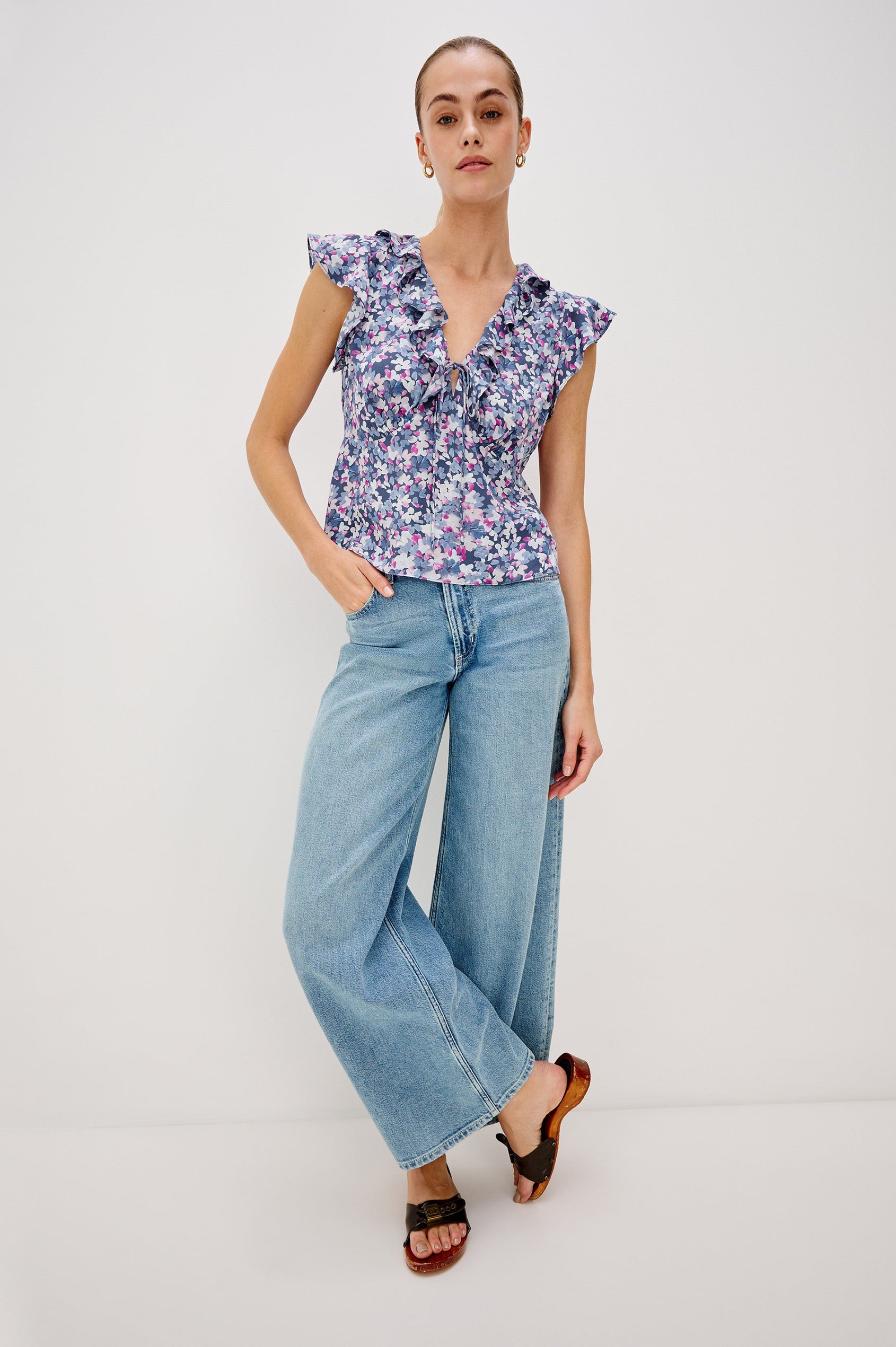 Carmine blouse, a chiffon top with ruffled cap sleeves, ruffles at the v-neckline, a front tie, and empire waist silhouette in Blue Blossom Colorway. Full Body Front View Shown on Model.
