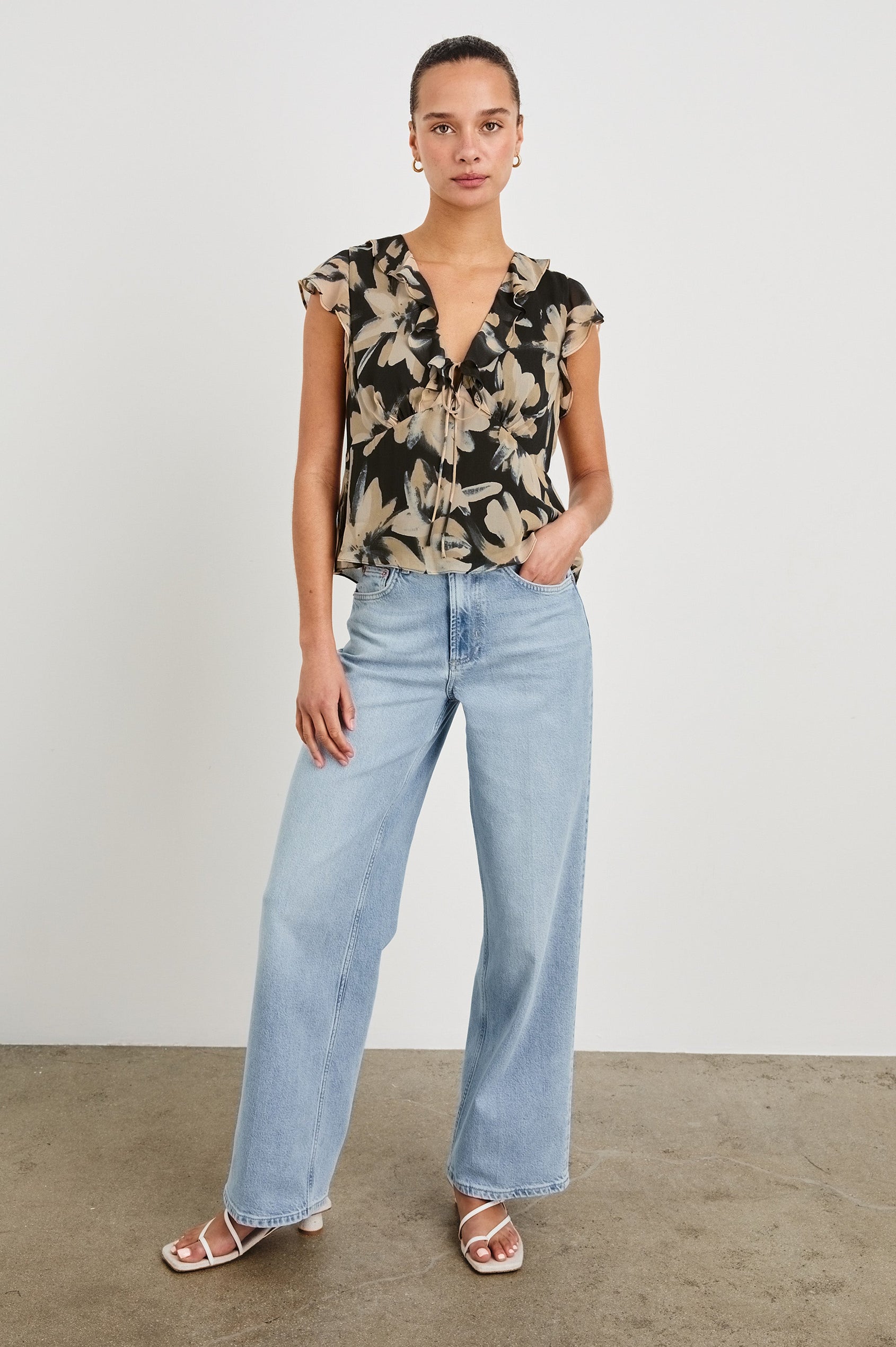 Full length view of model wearing our Carmine blouse, a chiffon top with flutter sleeves, ruffles at the v-neckline, a front tie, and empire waist silhouette. Styled with light washed denim pants.