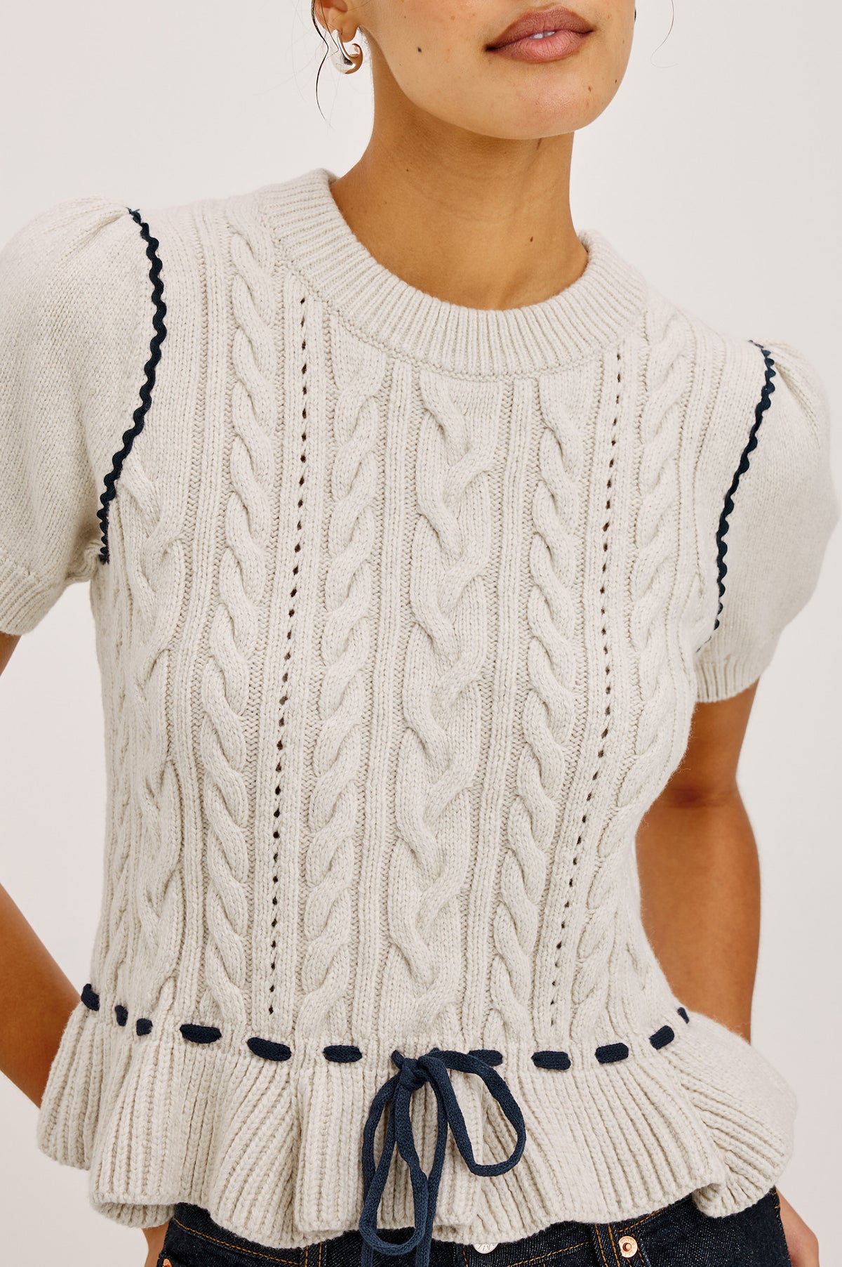 Chandi Sweater in Cable Knit With Short Puff Sleeves, Fitted SIlhouette, Soft Flared Peplum Hem, and Contrast Serpentine Trim and Interlocking Drawstring Ties in Heather Oat Colorway - Upper Body Front View Featured on Model