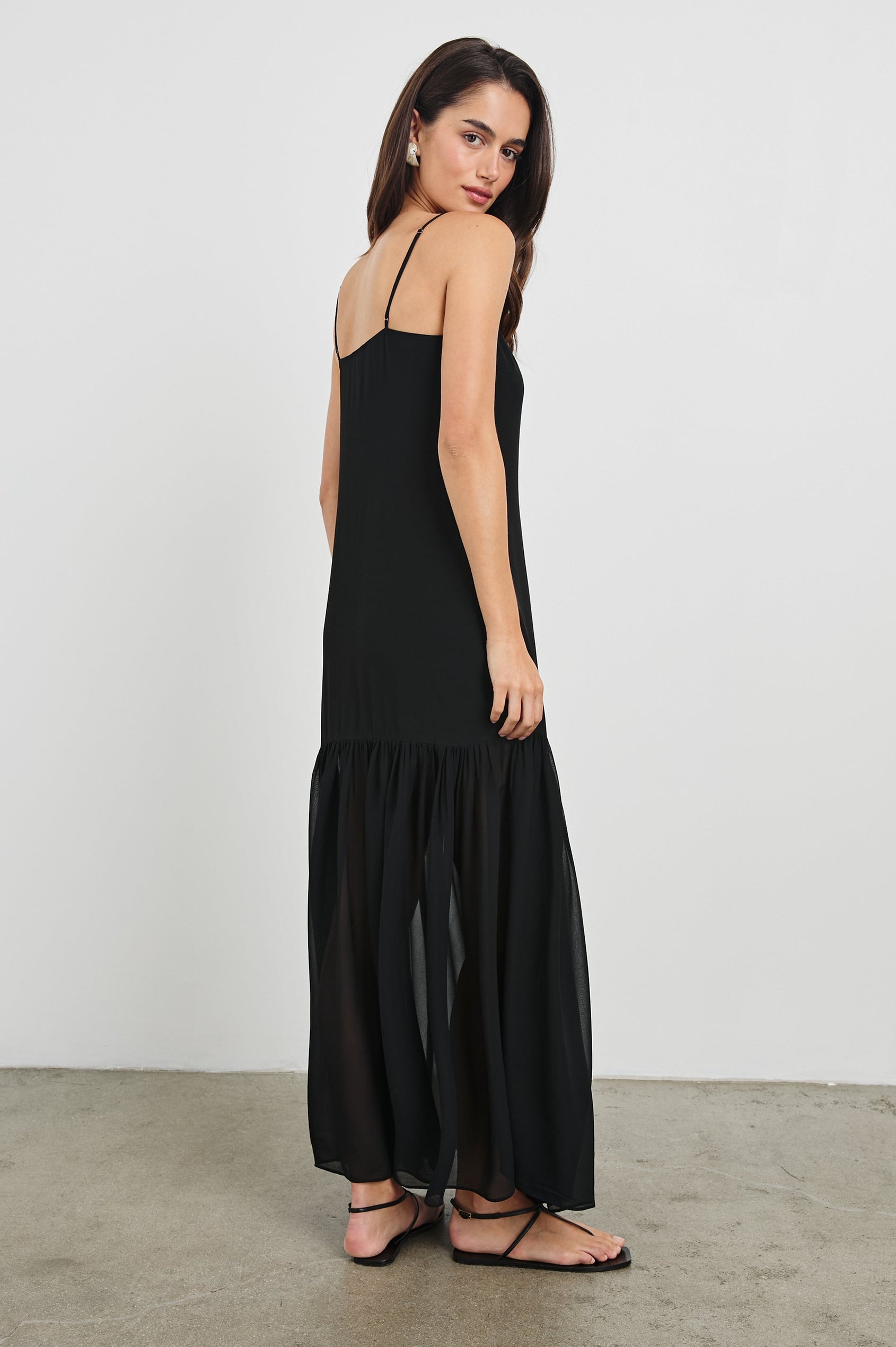 Chandra Maxi Tank Dress with Spaghetti Straps and a Contrast Semi Sheer Fabric Gathered Hem in Black Colorway. Full Body Back View Shown on Model.