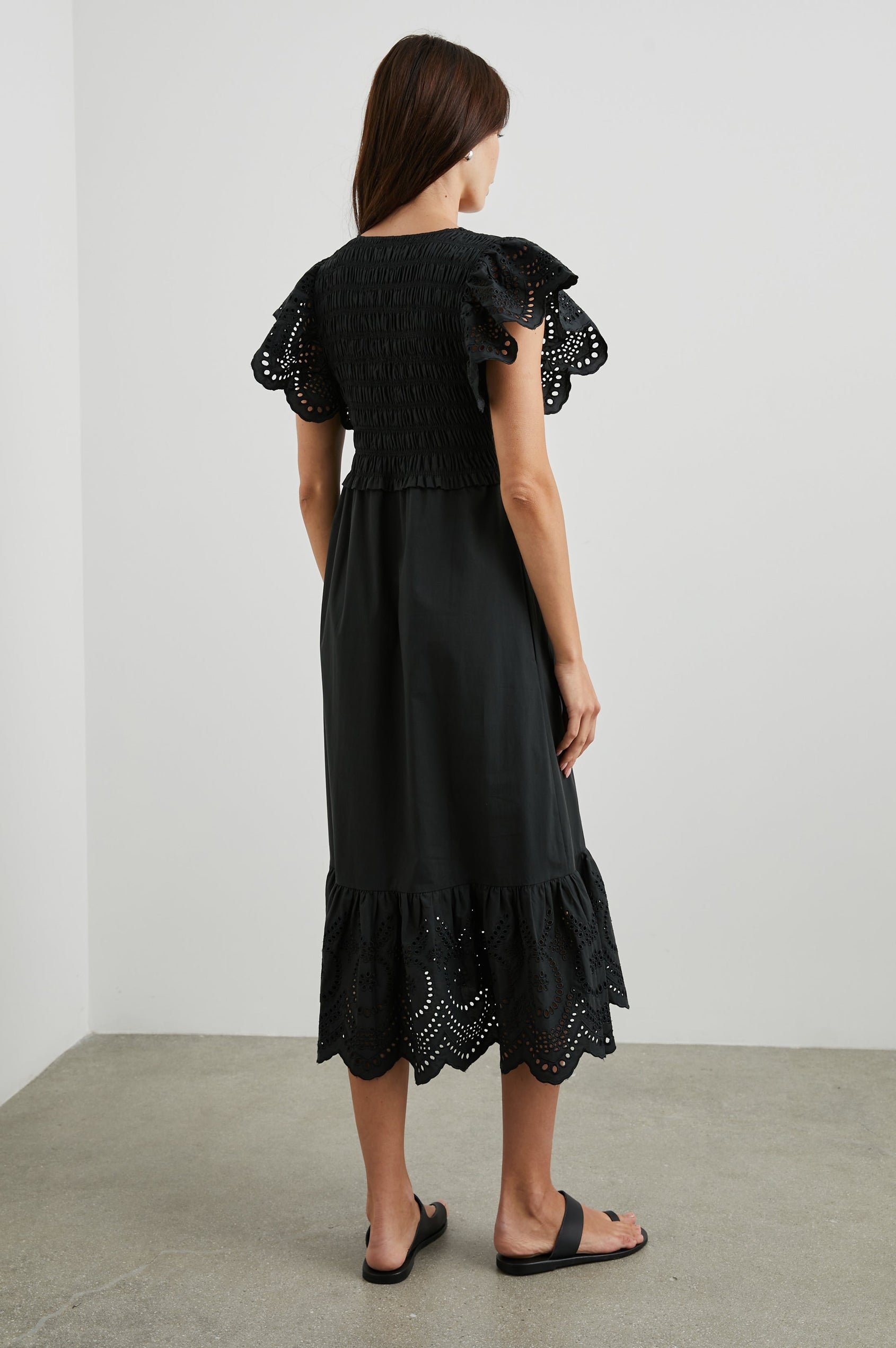 Clementine Midi Dress with a Deep V Neckline, Flutter Sleeves, Smocked Top, Two Side Pockets and eyelets on the Sleeves and Hem in Black Eyelet Colorway- Back Full Body View Featured on Model