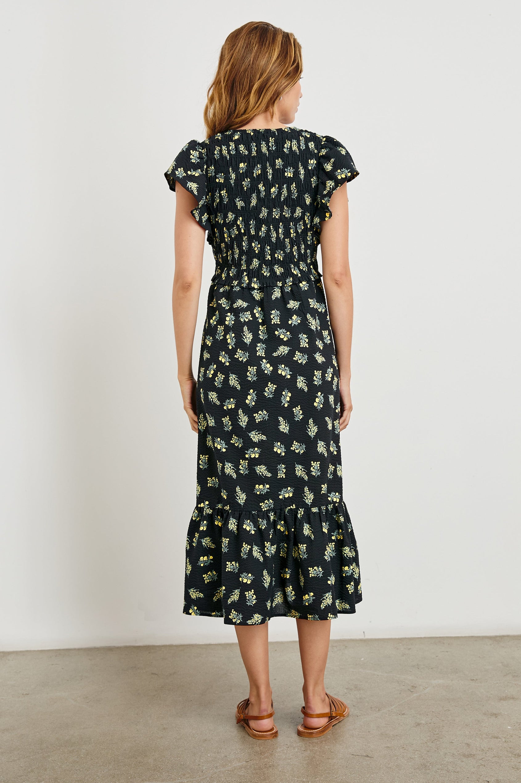 Clementine Midi Dress with a Deep V Neckline, Flutter Sleeves, Smocked Top and Two Side Pockets in Dark Amaryllis Colorway, Yellow and Green flowers on Black - Back Full Body View Featured on Model