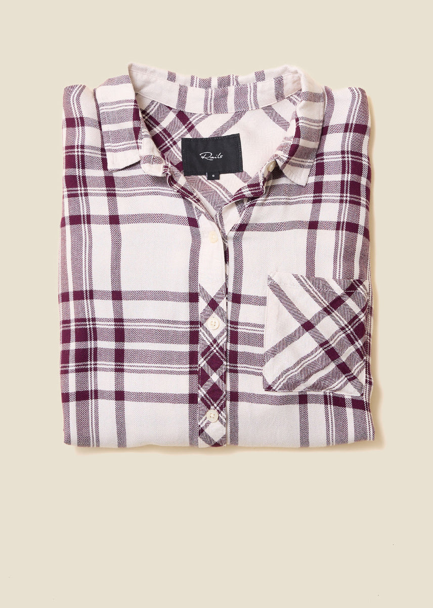 A folded long-sleeve plaid button-up shirt in white with purple and gray stripes, featuring a chest pocket and a black brand label, set against a solid beige background.