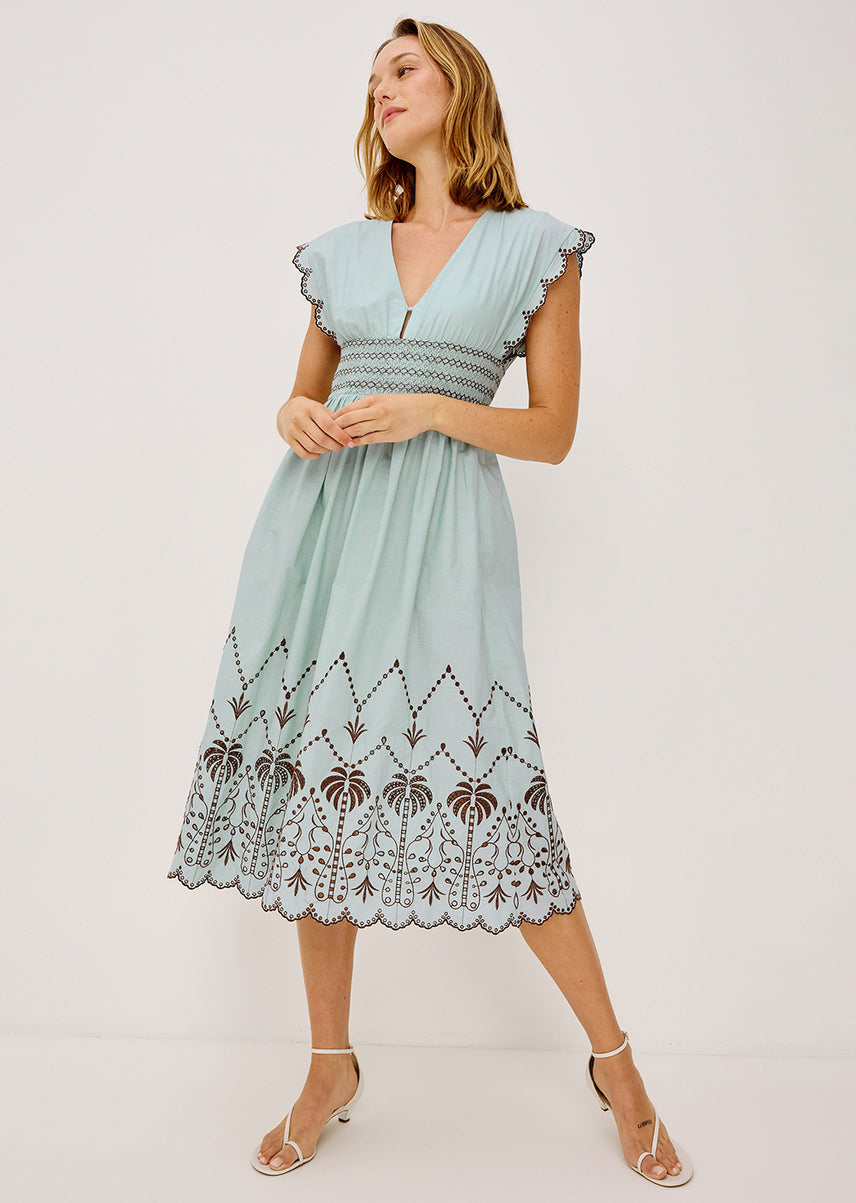 A woman modeling a light blue midi dress with ruffled cap sleeves, a smocked waist, and a brown embroidered floral border near the hem, photographed against a plain light background.