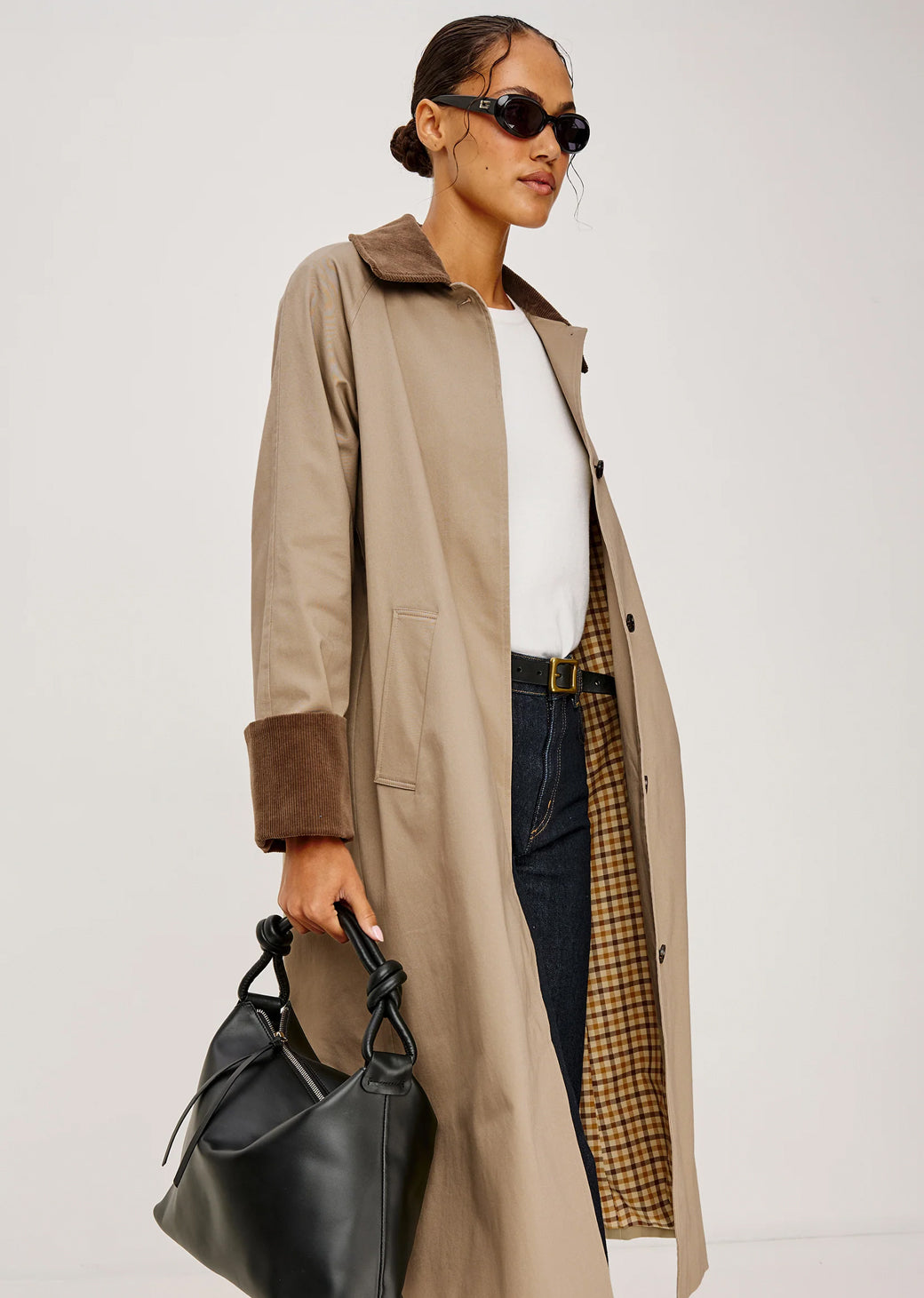 Woman wearing a beige trench coat holding a black handbag against a white background.