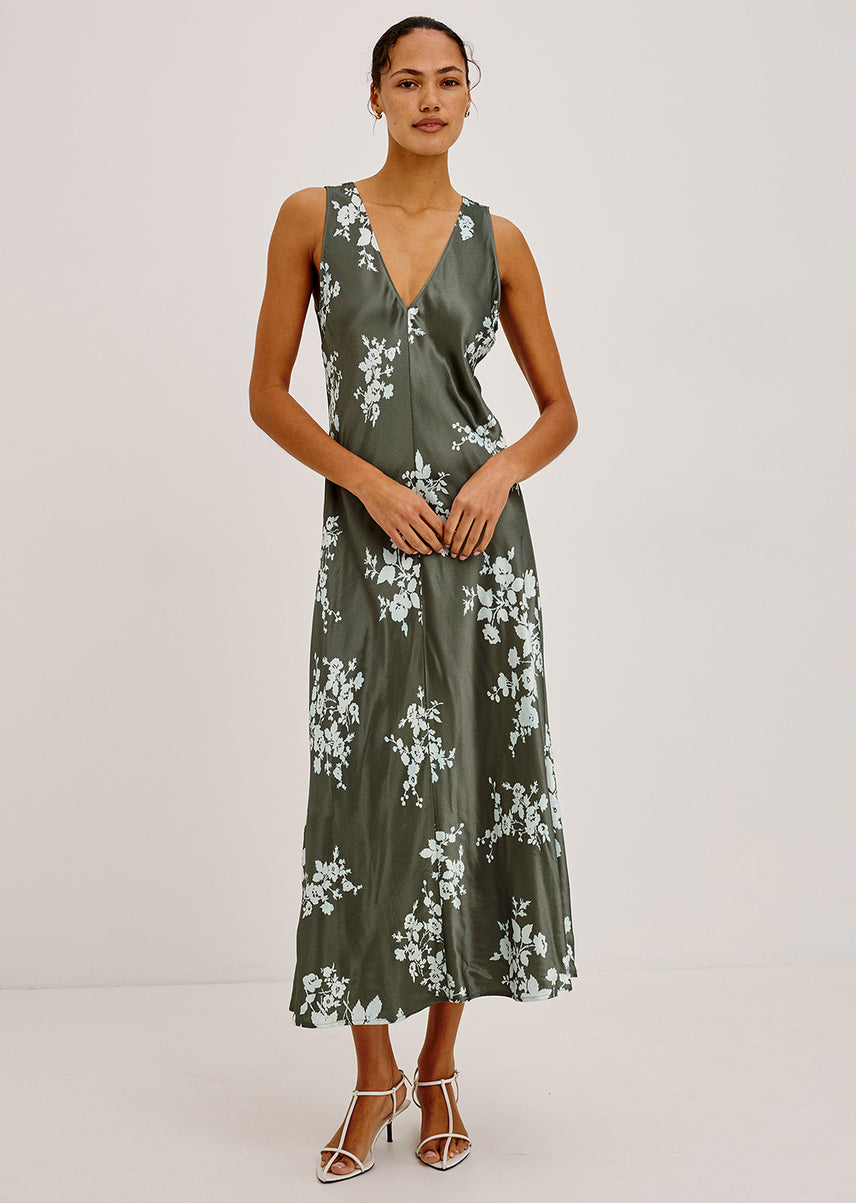 A woman modeling a sleeveless olive green midi dress with a white floral print and a wrap-style V-neckline, paired with white strappy high-heeled sandals against a plain light gray studio background.