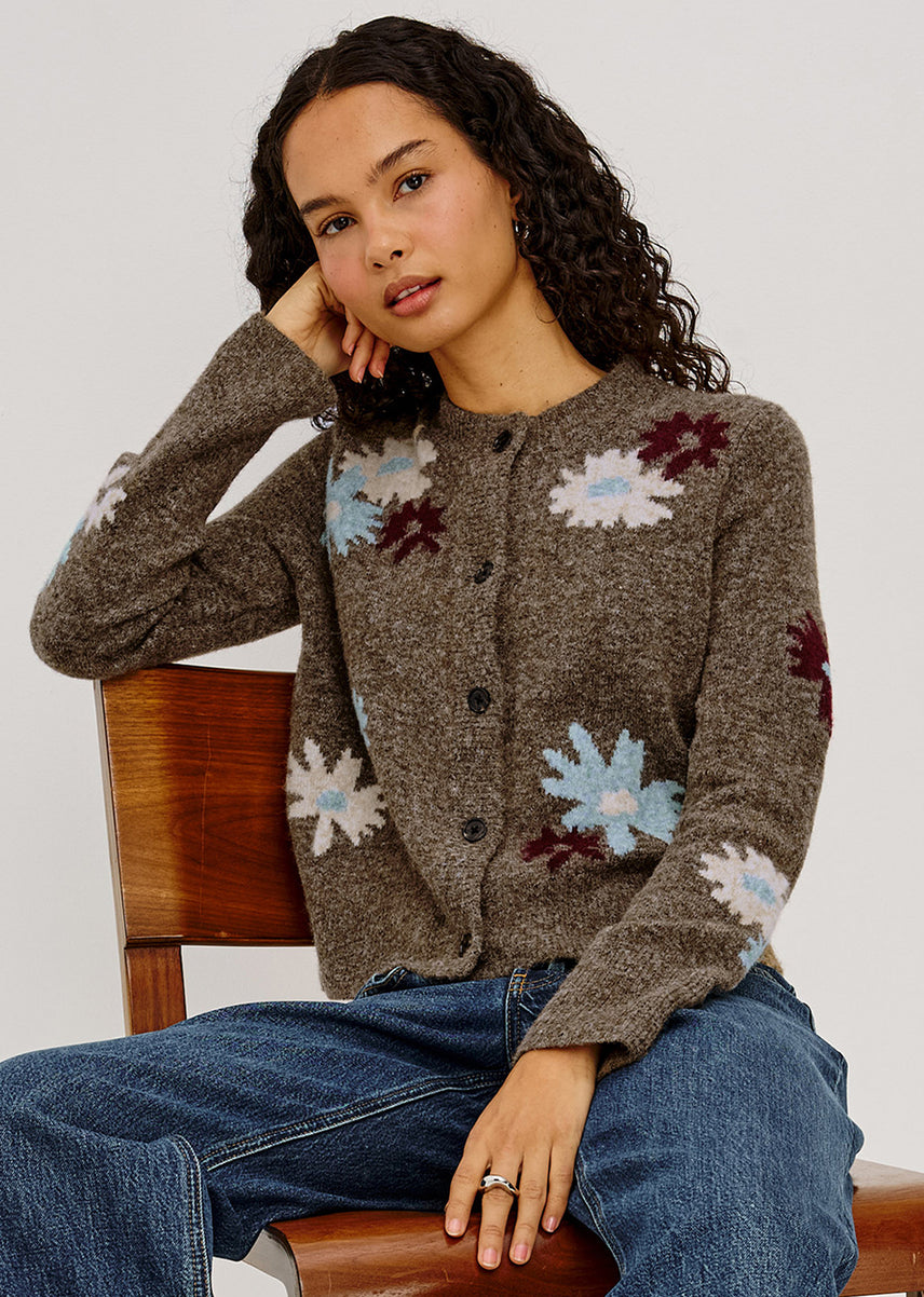 A woman with curly dark hair sits on a wooden chair wearing a brown knit cardigan with light blue, white, and burgundy floral motifs, paired with blue jeans against a plain light gray background.