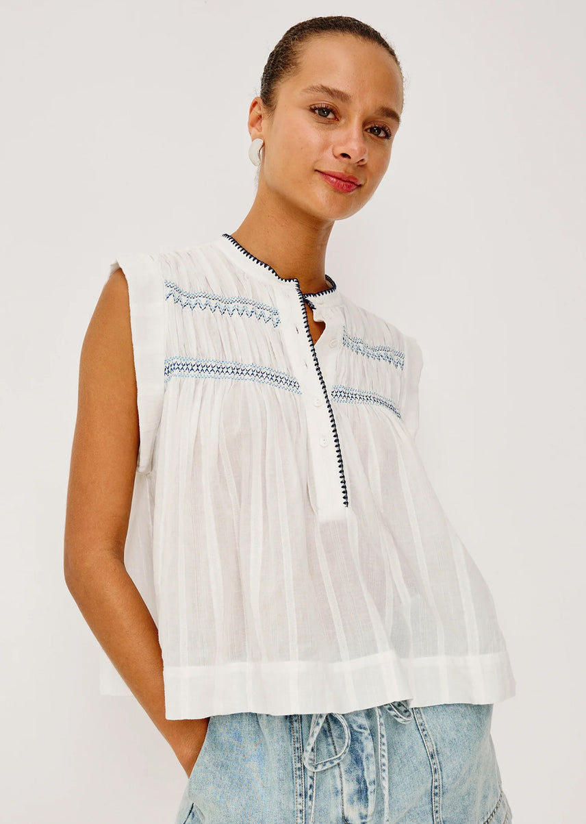 Woman wearing a white sleeveless top with blue embroidery on a plain background