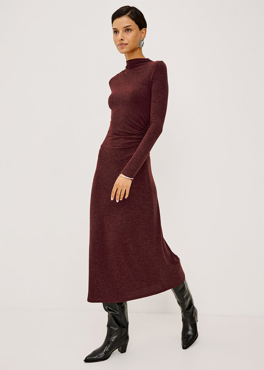A woman modeling a long-sleeve, mock-neck midi dress in a deep burgundy color, paired with black leather ankle boots, against a plain light gray studio background.