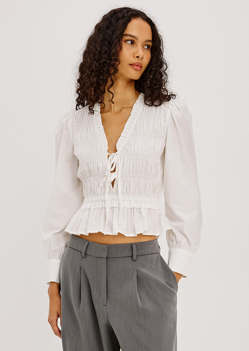 A woman wearing a white long-sleeve smocked blouse with a tie-front detail and a ruffled peplum hem, paired with gray trousers, against a light gray studio background.
