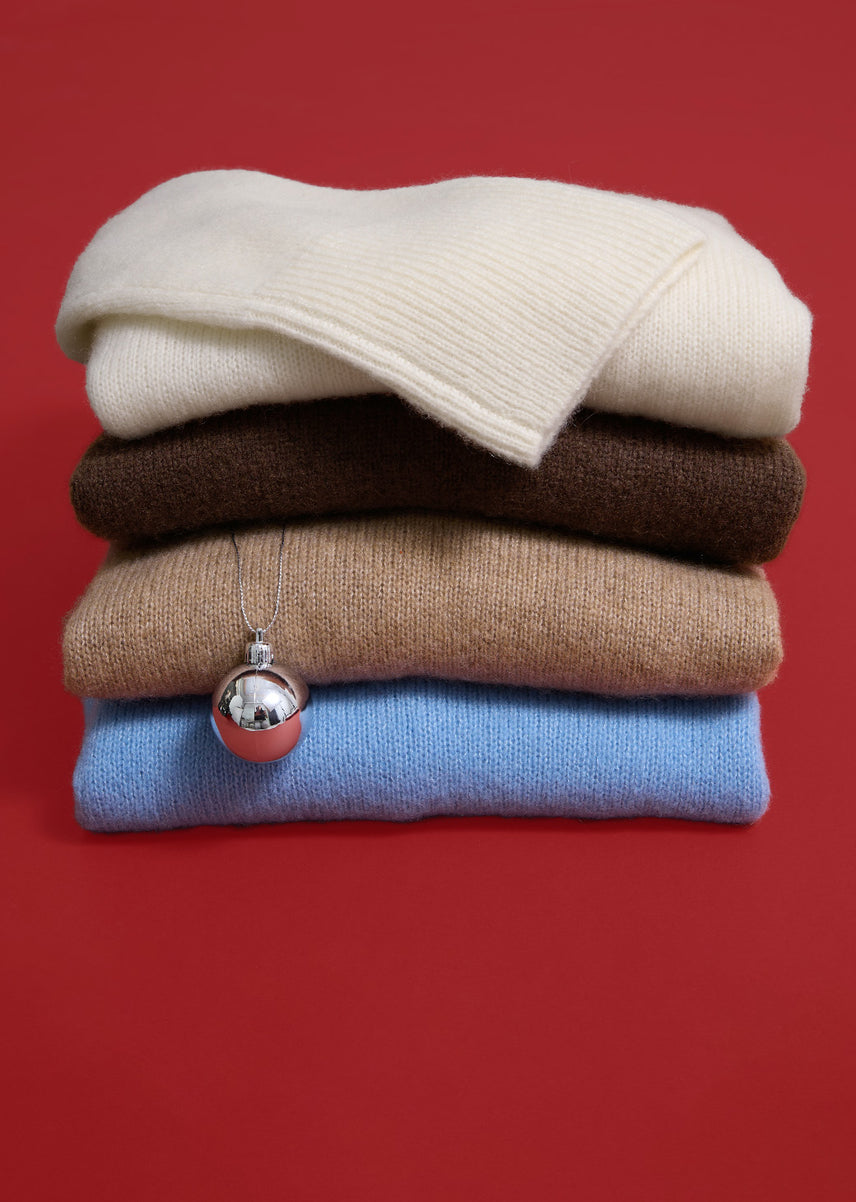 A stack of four folded knit sweaters in cream, brown, beige, and light blue, with a silver ornament pendant resting on the front, set against a solid red background.
