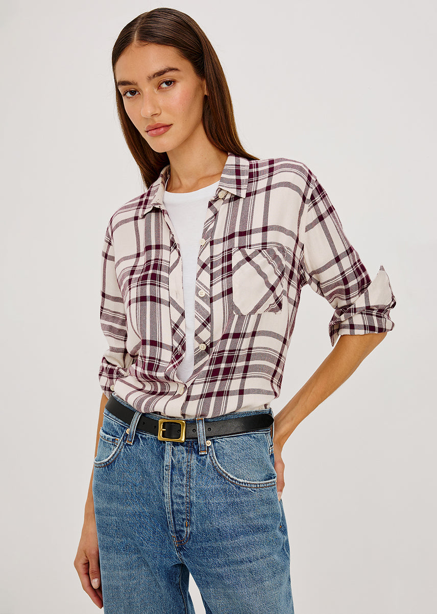 A woman modeling a long-sleeve plaid button-up shirt with rolled sleeves, tucked into a white t-shirt and paired with blue jeans and a black belt, against a plain light gray background.