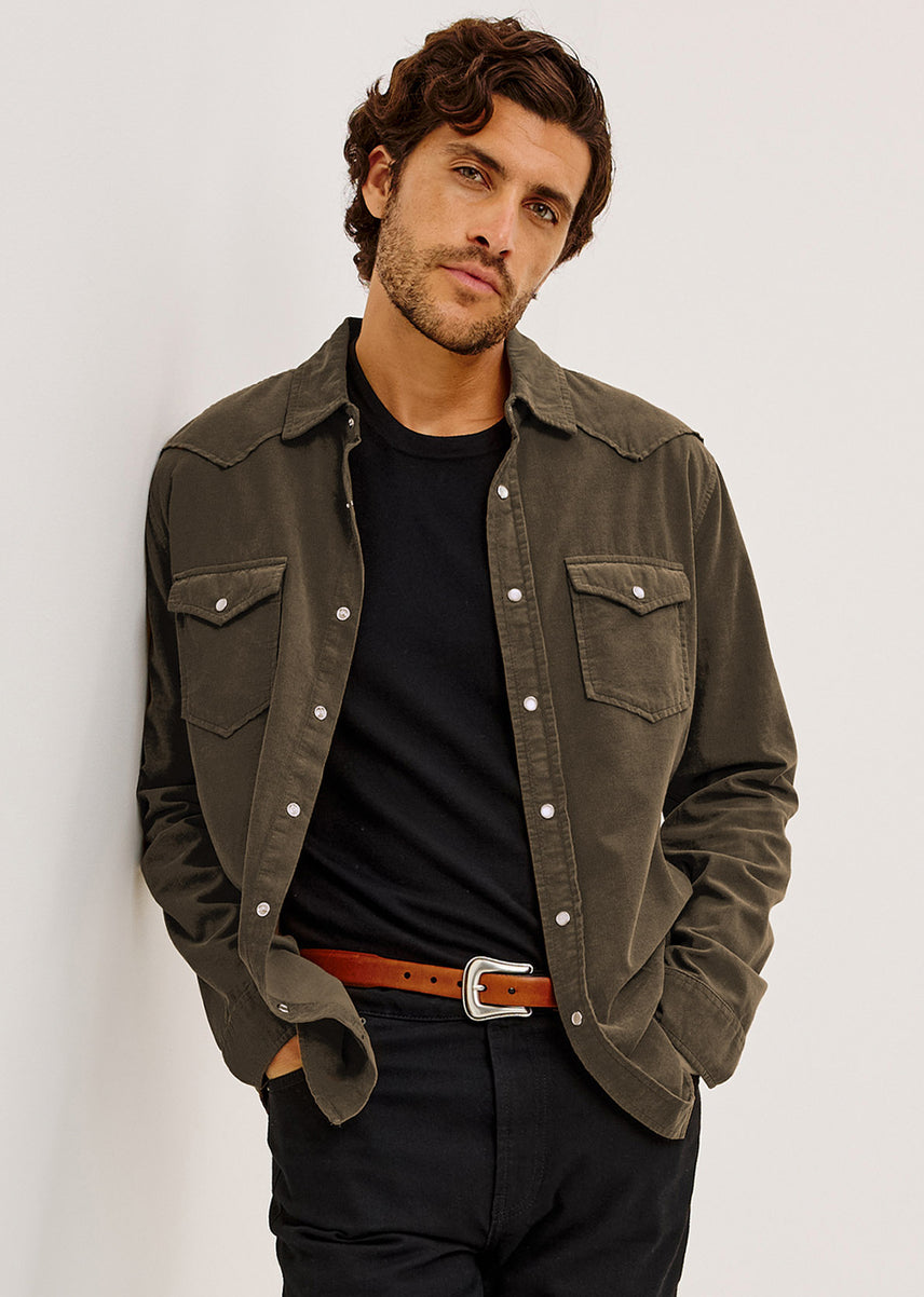 A man wearing an olive green western-style button-up shirt jacket over a black t-shirt, paired with a brown leather belt and dark trousers, posing against a light gray wall.