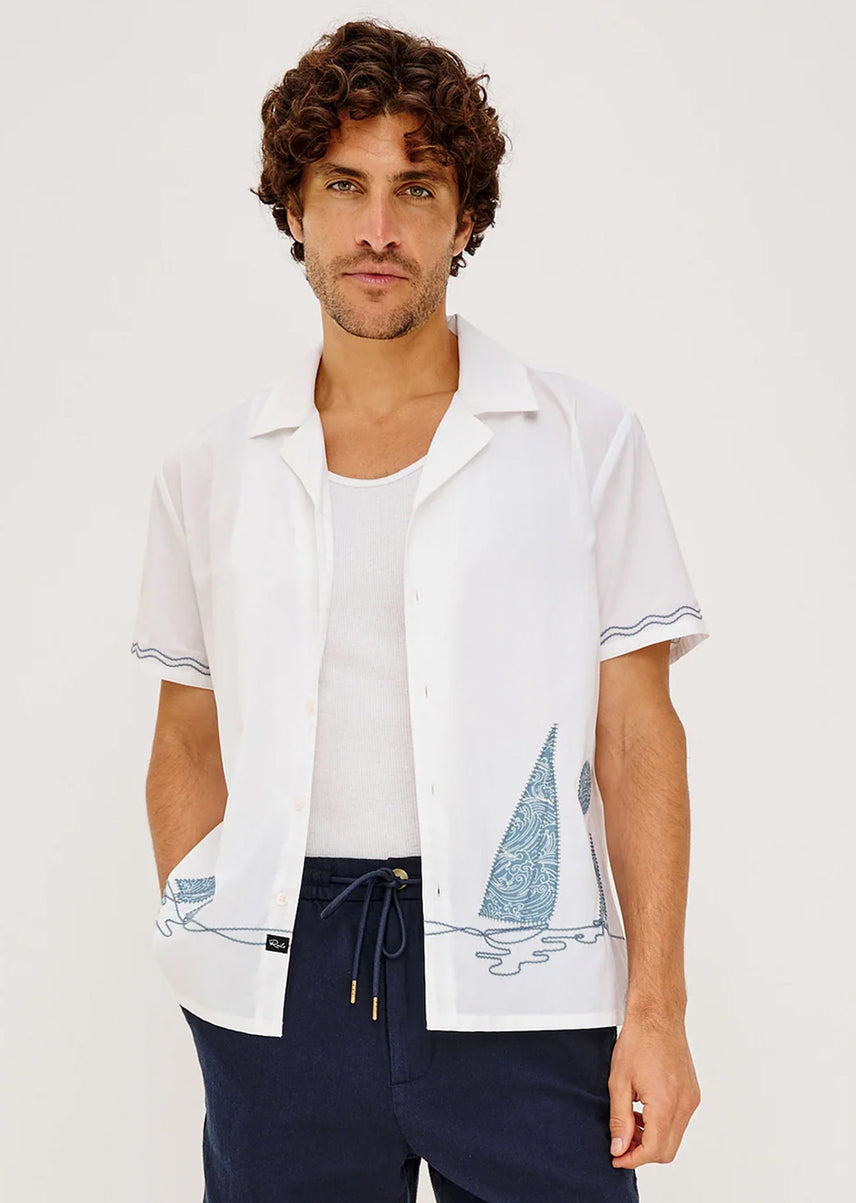 A man wearing a white short-sleeve button-up shirt with blue sailboat and wave prints, layered over a white t-shirt and navy pants, against a plain white background.