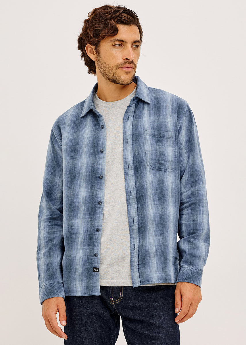 A man wearing a blue and gray plaid button-down shirt over a light gray t-shirt and dark jeans, standing against a plain light gray studio background.
