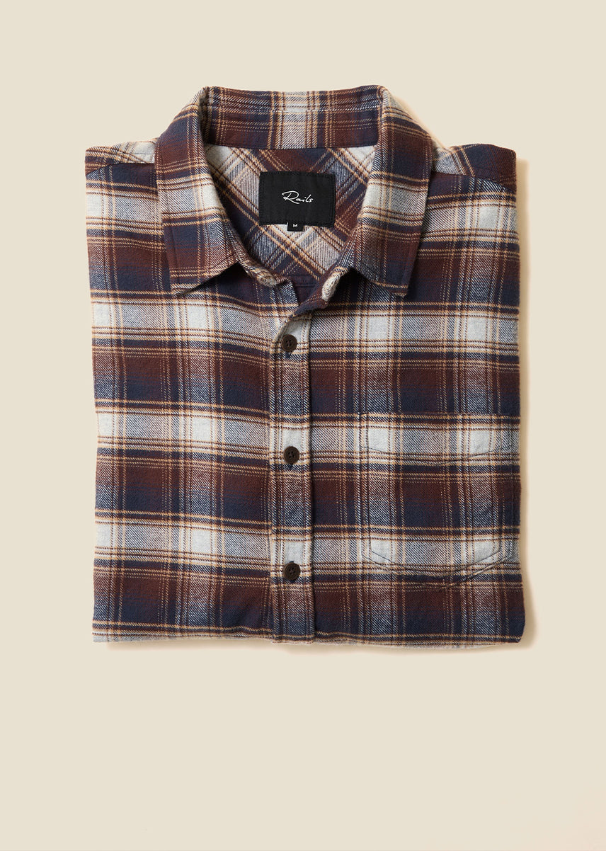 A folded long-sleeve plaid button-up shirt with a collar, displayed flat against a solid beige background. The shirt features a plaid pattern of dark brown, navy blue, white, and thin orange stripes.