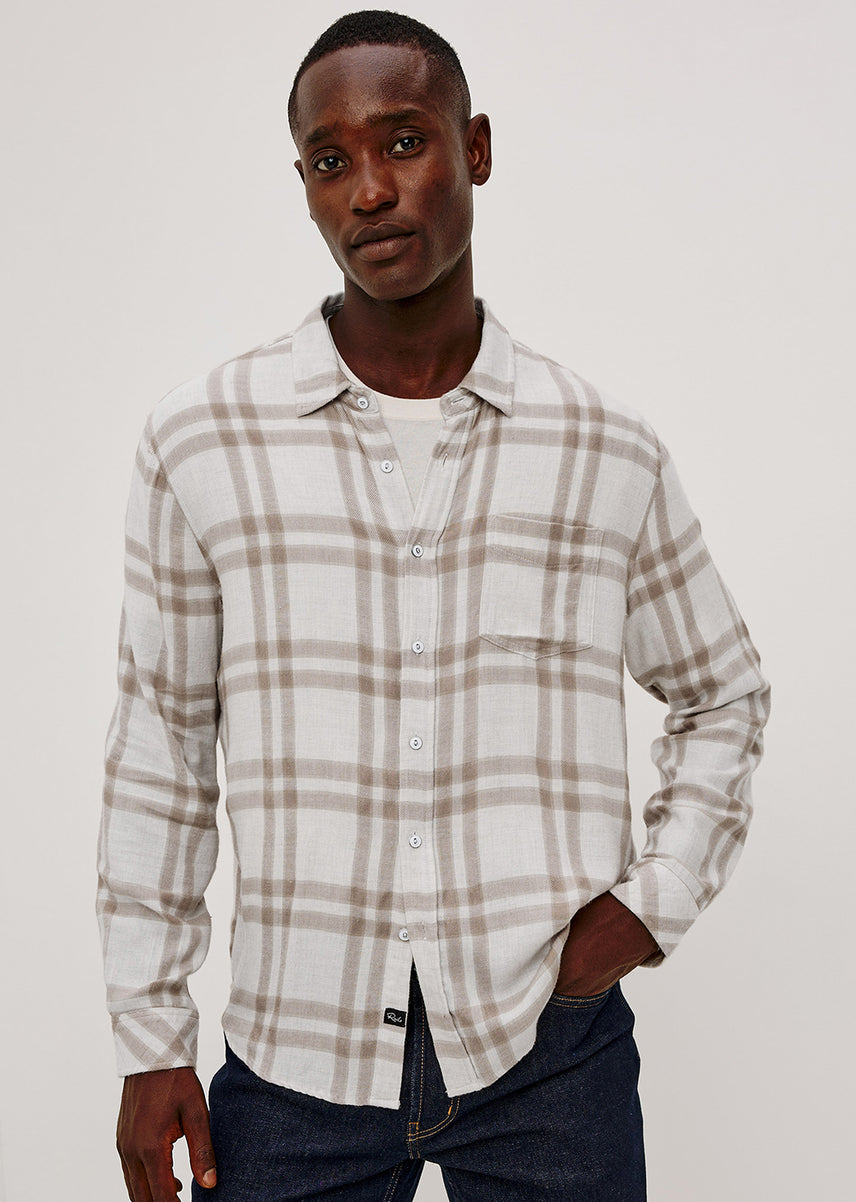 A man wearing a long-sleeve button-up plaid shirt in beige and white, paired with dark blue jeans, photographed against a plain light gray studio background.