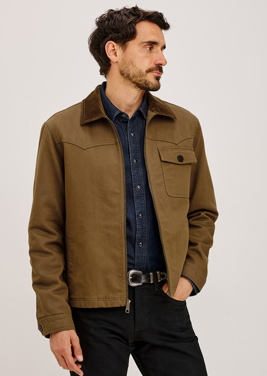 A man wearing a brown canvas utility jacket over a dark blue button-down shirt and black pants, standing against a light gray studio background.