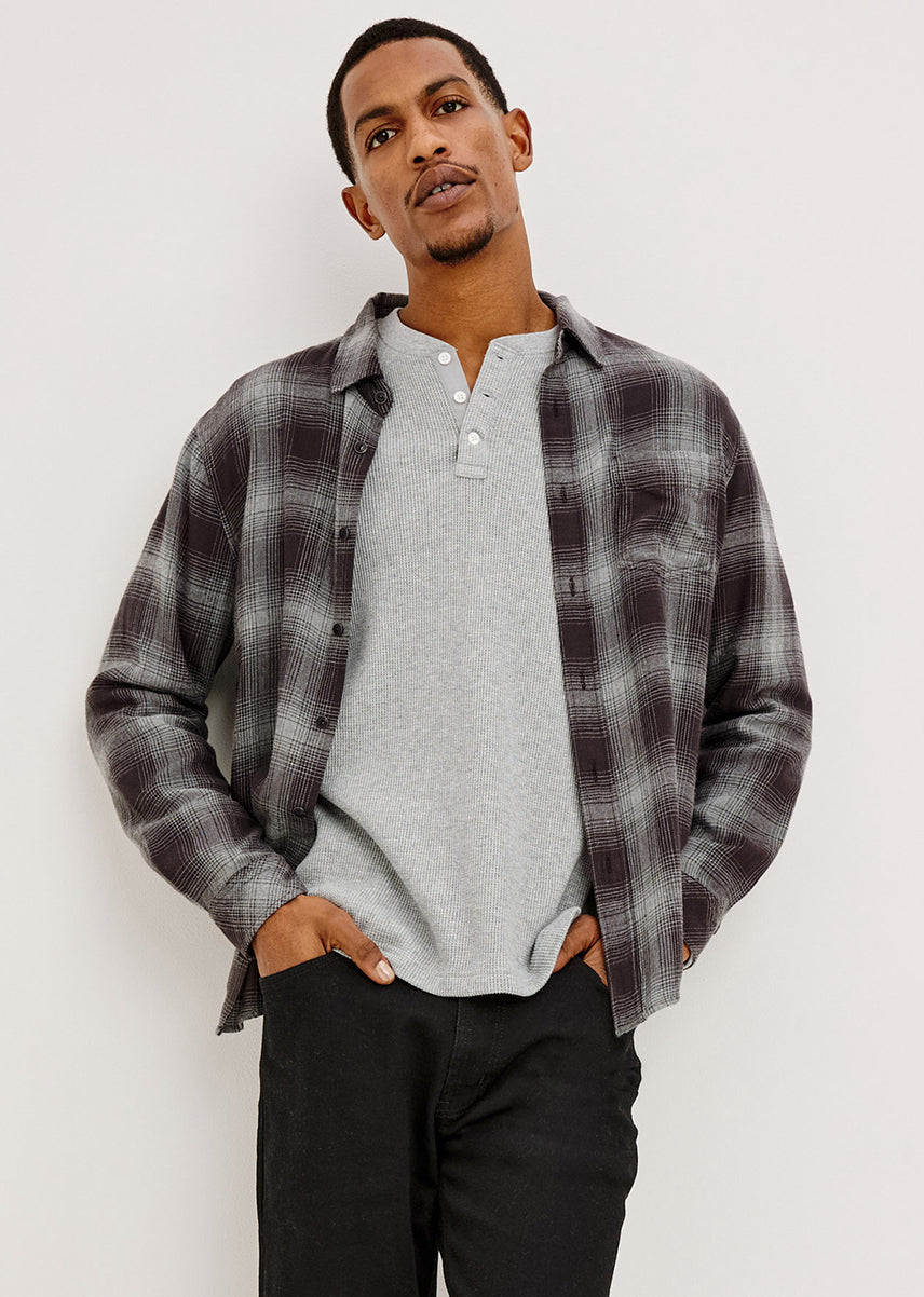 A man wearing a dark gray and black plaid button-down shirt over a light gray henley t-shirt and black pants, standing against a plain off-white wall.