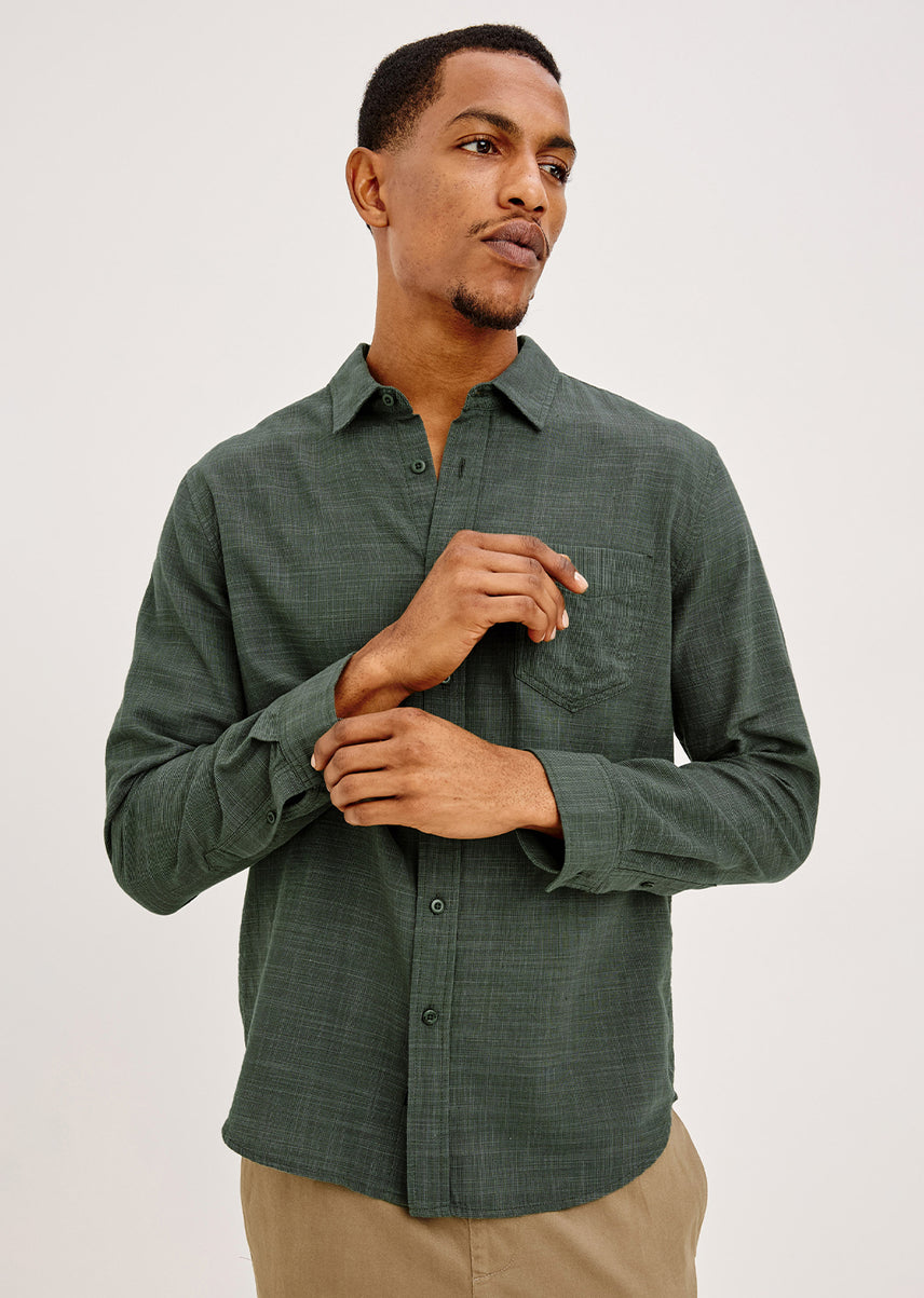 A man wearing a dark green long-sleeve button-up shirt with a subtle textured pattern, paired with khaki pants, photographed against a light gray studio background.