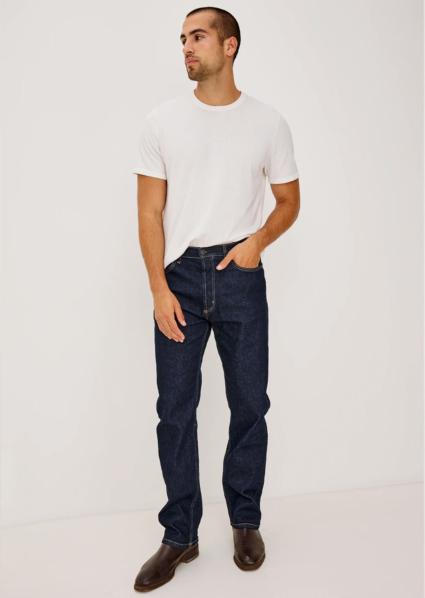 A man wearing a white crew-neck t-shirt, dark blue straight-leg jeans, and brown leather shoes, standing against a plain white studio background.