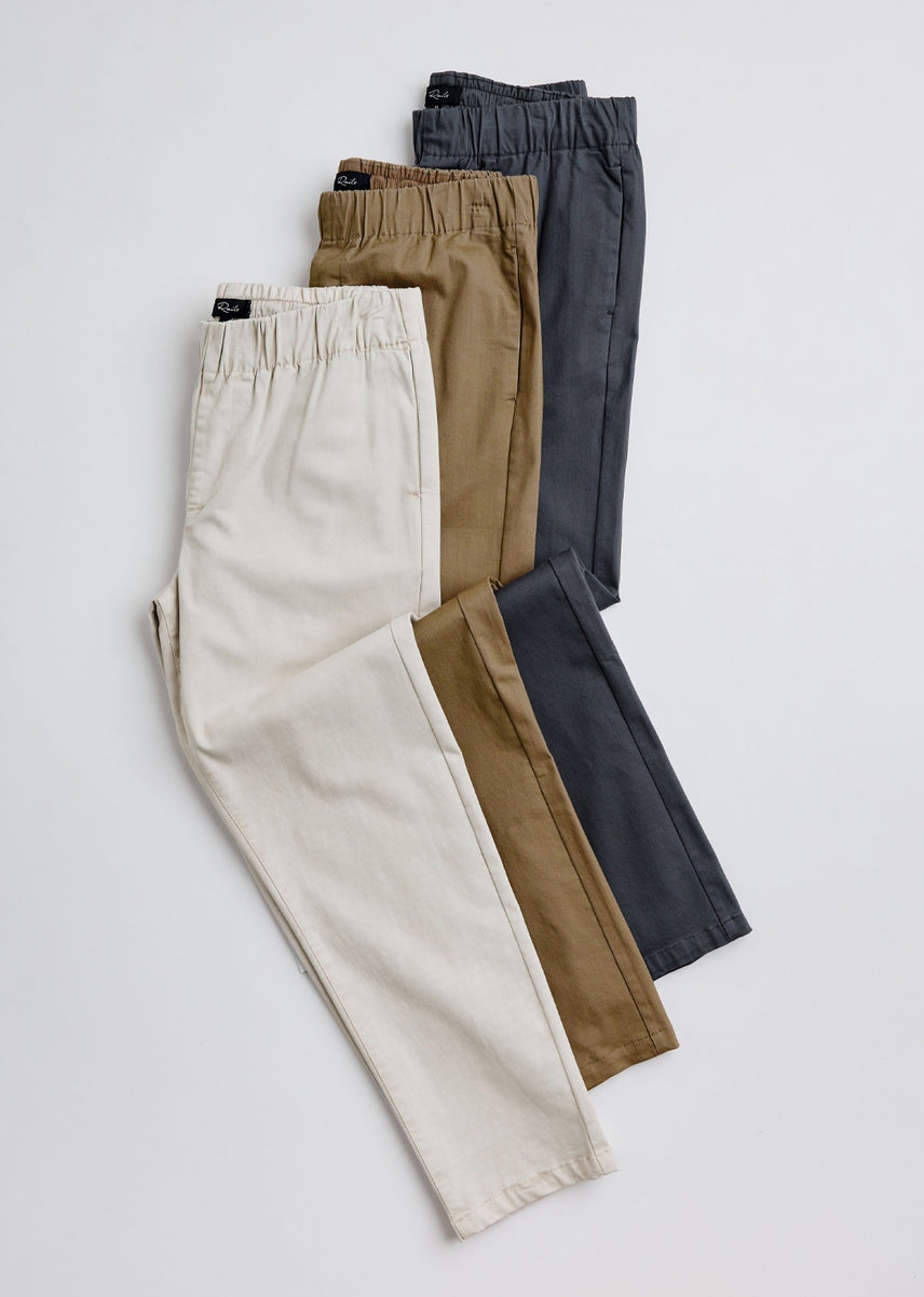 Three pairs of men's elastic-waist trousers in cream, tan, brown, and charcoal gray, arranged in a fan shape on a light gray background.