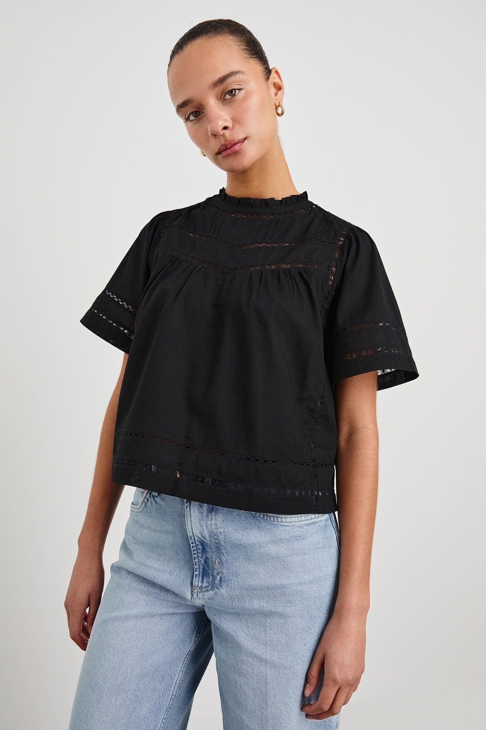 Front view of female model wearing our Daisy Top in black. This is a crewneck baby doll blouse, with ruffles at the collar. Styled with light wash denim and a simple belt.