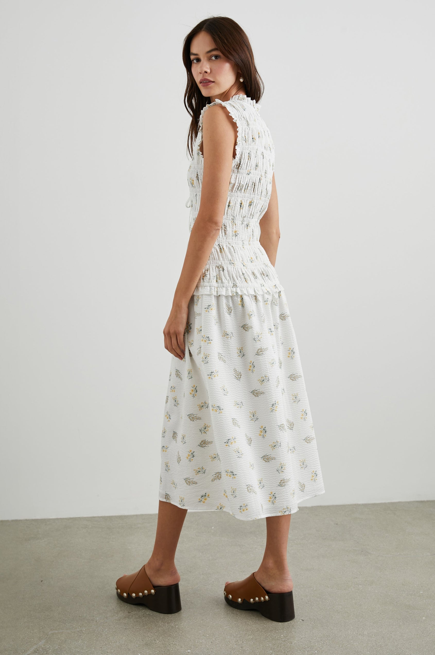 Dio Sleeveless Midi Dress with a Deep V Neck with a Two Tie Closure, a Shirred Bodice and Ruffled Trim on the Bodice in Amaryllis Colorway, Small Yellow and Green Flowers on White - Side Full Body View Featured on Model 