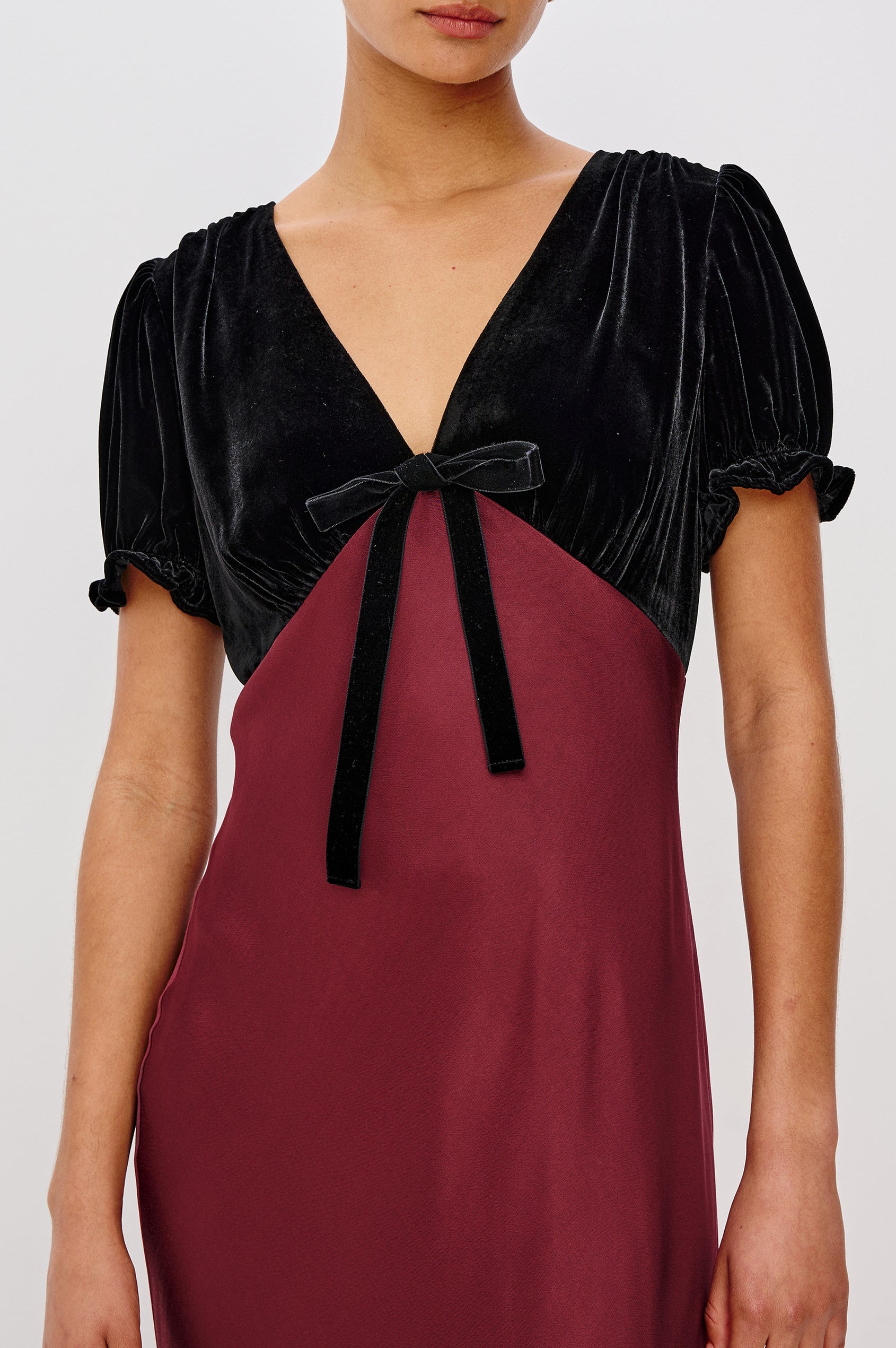 A garnet velvet maxi dress with a fitted V-neck bodice and adjustable center-bust ties. The bias-cut skirt drapes elegantly to ankle length, finished with a back zipper closure. Close Up View on Model.