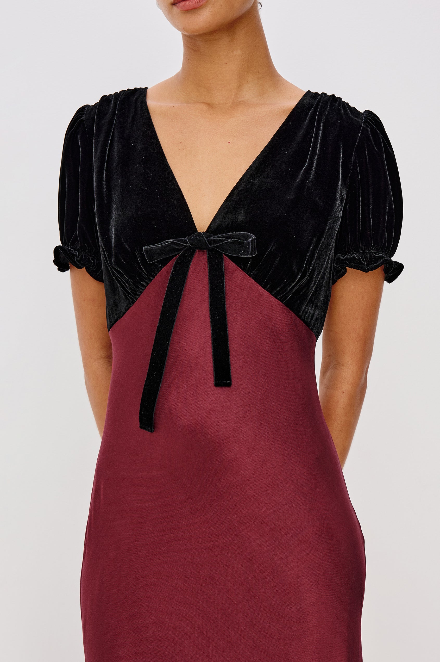 A garnet velvet maxi dress with a fitted V-neck bodice and adjustable center-bust ties. The bias-cut skirt drapes elegantly to ankle length, finished with a back zipper closure. Close Up View on Model.