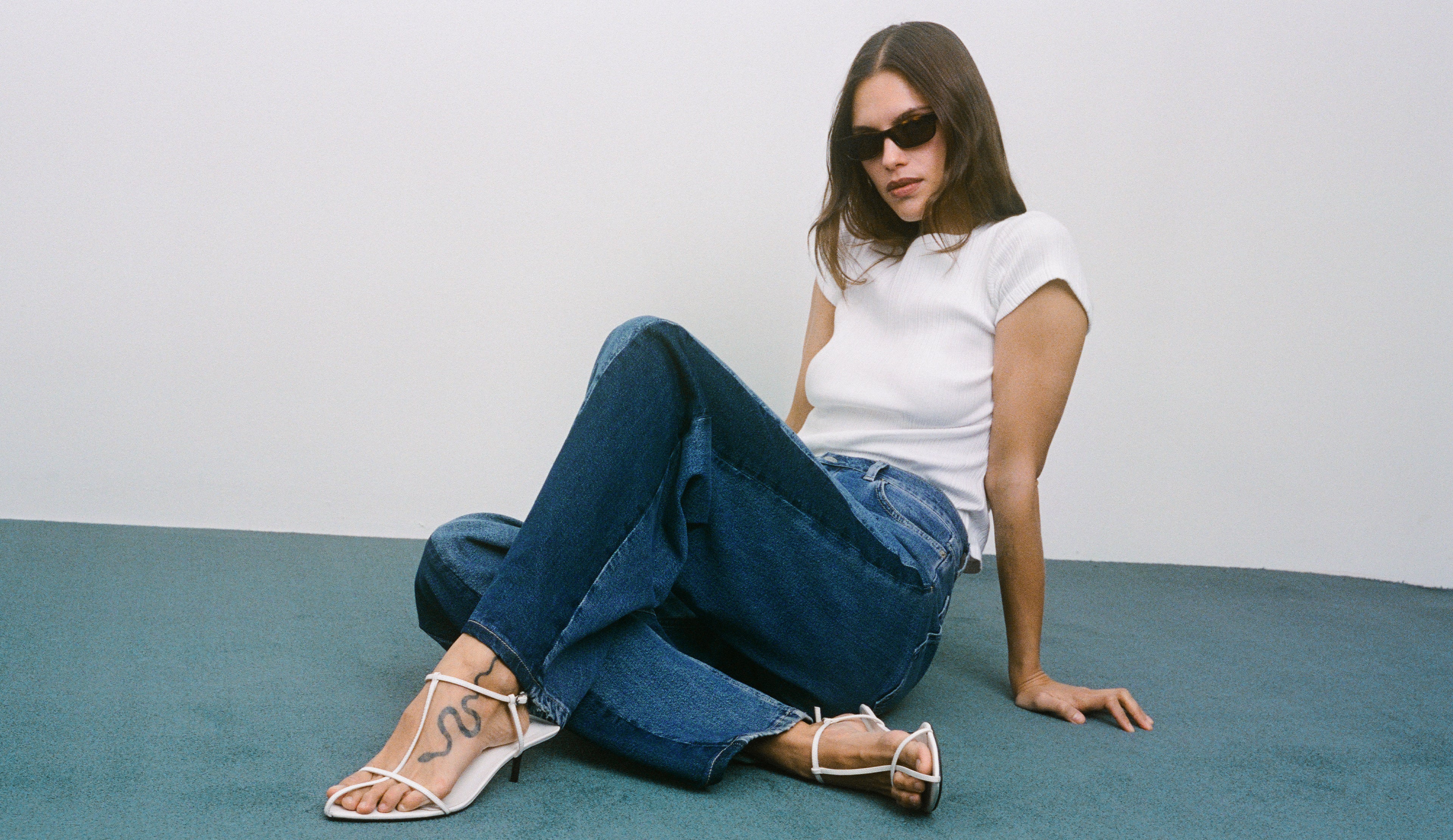 Woman sitting on a blue carpet wearing a white t-shirt, blue jeans, and white heels.