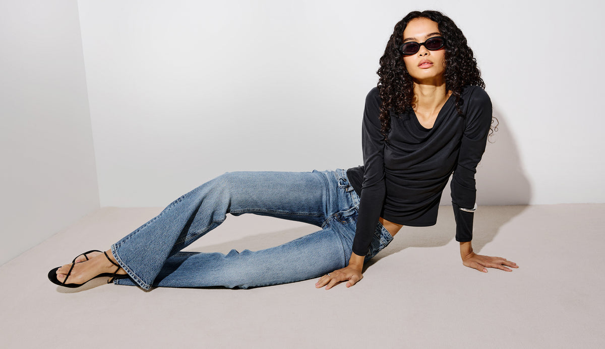 A model wearing a black long-sleeve cowl-neck top, light-wash blue flare jeans, and black strappy heels, posing on a minimalist light gray studio floor and wall.