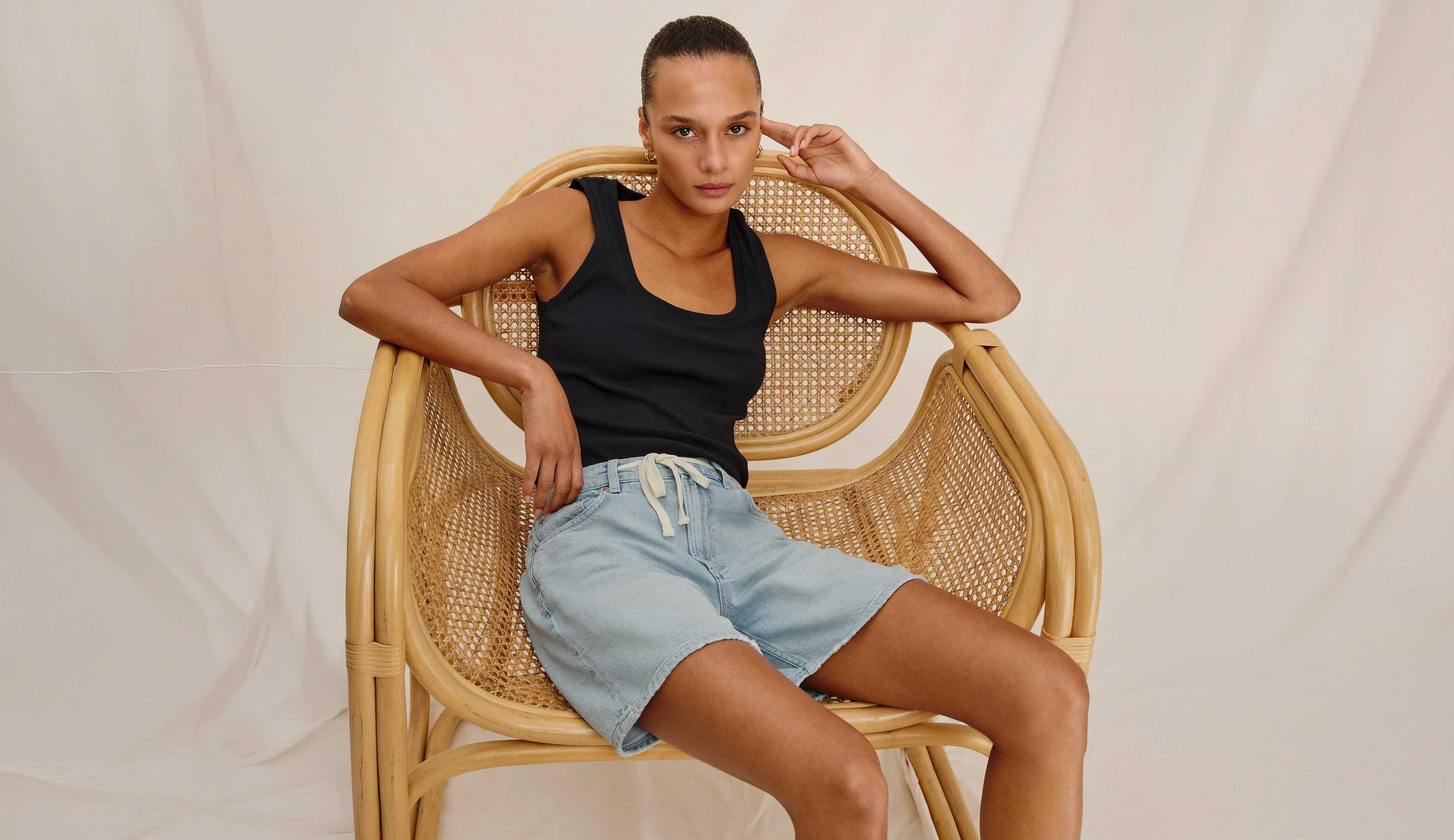Woman sitting on a wicker chair wearing a black tank top and light blue shorts against a plain background