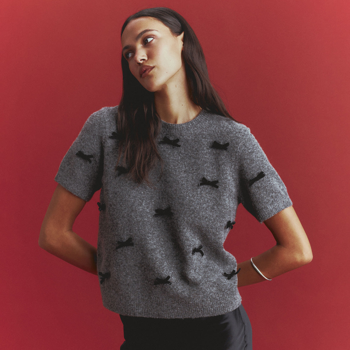 Woman in a gray short-sleeve knit sweater scattered with small black velvet bows, worn with a black skirt and a silver bangle, against a red backdrop.