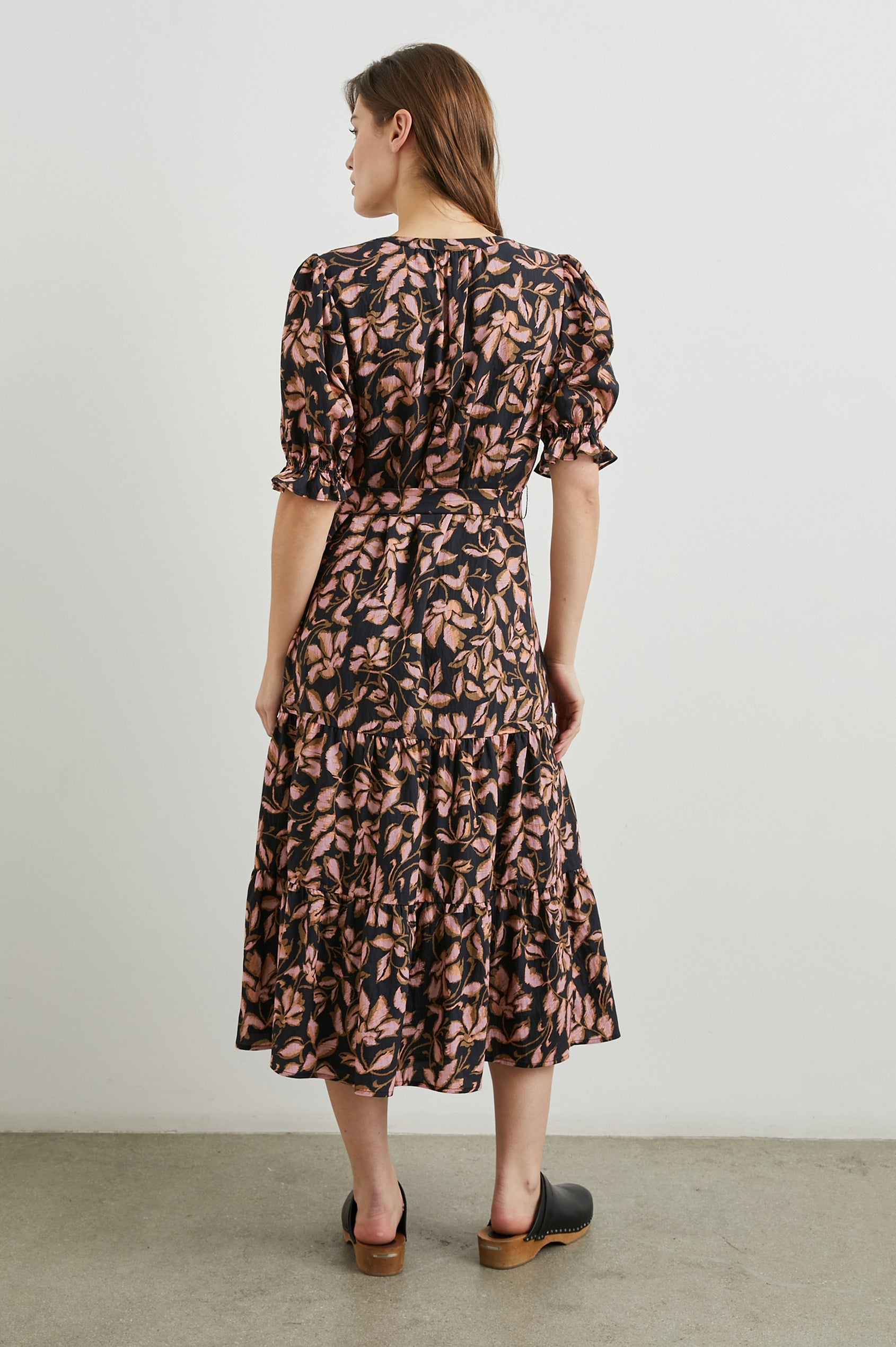 Eliana Midi Dress with Elbow-Length Sleeves with Shirred Cuffs, a V Neck, Buttons Down the Bodice, a Fabric Tie at the Waist  and a Tiered Skirt in Jasmine Colorway, a Floral Pattern in Pink and Brown on Black - Back Full Body View Featured on Model