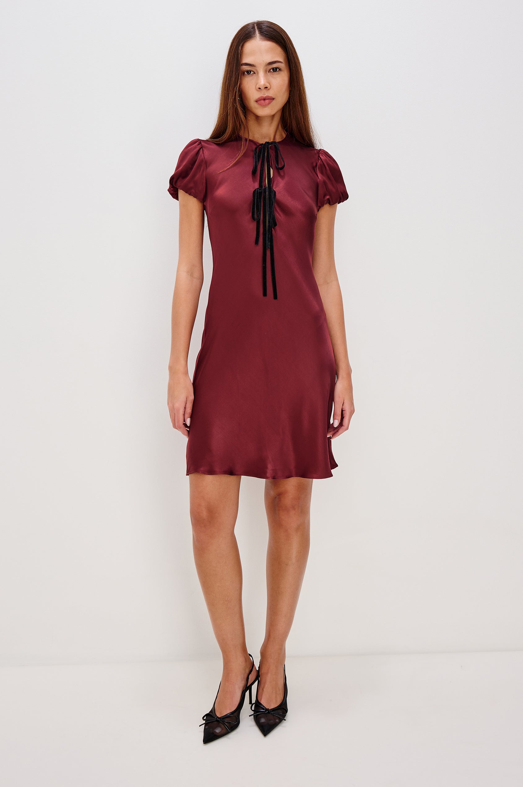 A garnet satin-back crepe midi dress cut on the bias. The dress features short puff sleeves, a deep V-neckline with velvet bow self-ties, and a fluid, flattering silhouette. Front View Featured on Model.