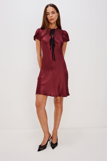 A garnet satin-back crepe midi dress cut on the bias. The dress features short puff sleeves, a deep V-neckline with velvet bow self-ties, and a fluid, flattering silhouette. Front View Featured on Model.