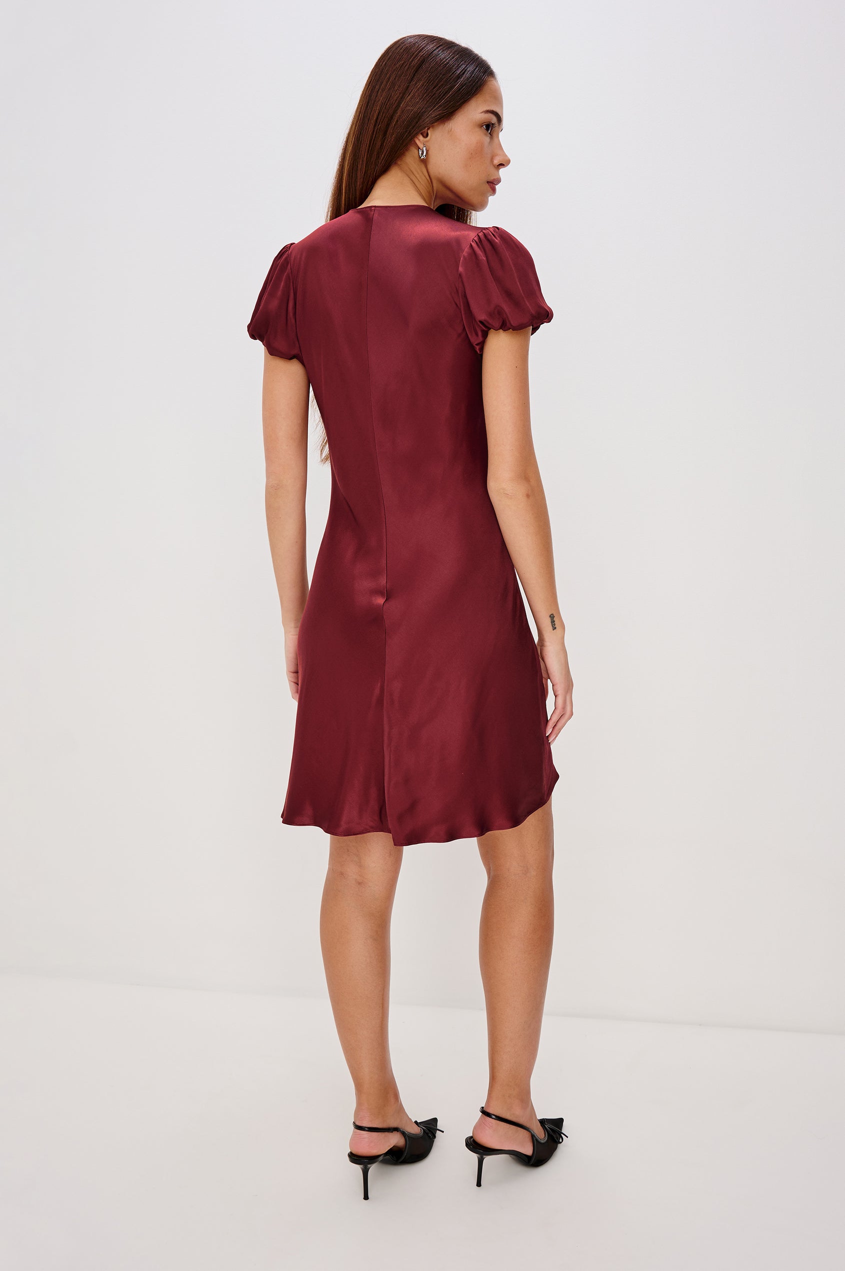 A garnet satin-back crepe midi dress cut on the bias. The dress features short puff sleeves, a deep V-neckline with velvet bow self-ties, and a fluid, flattering silhouette. Back View Featured on Model.