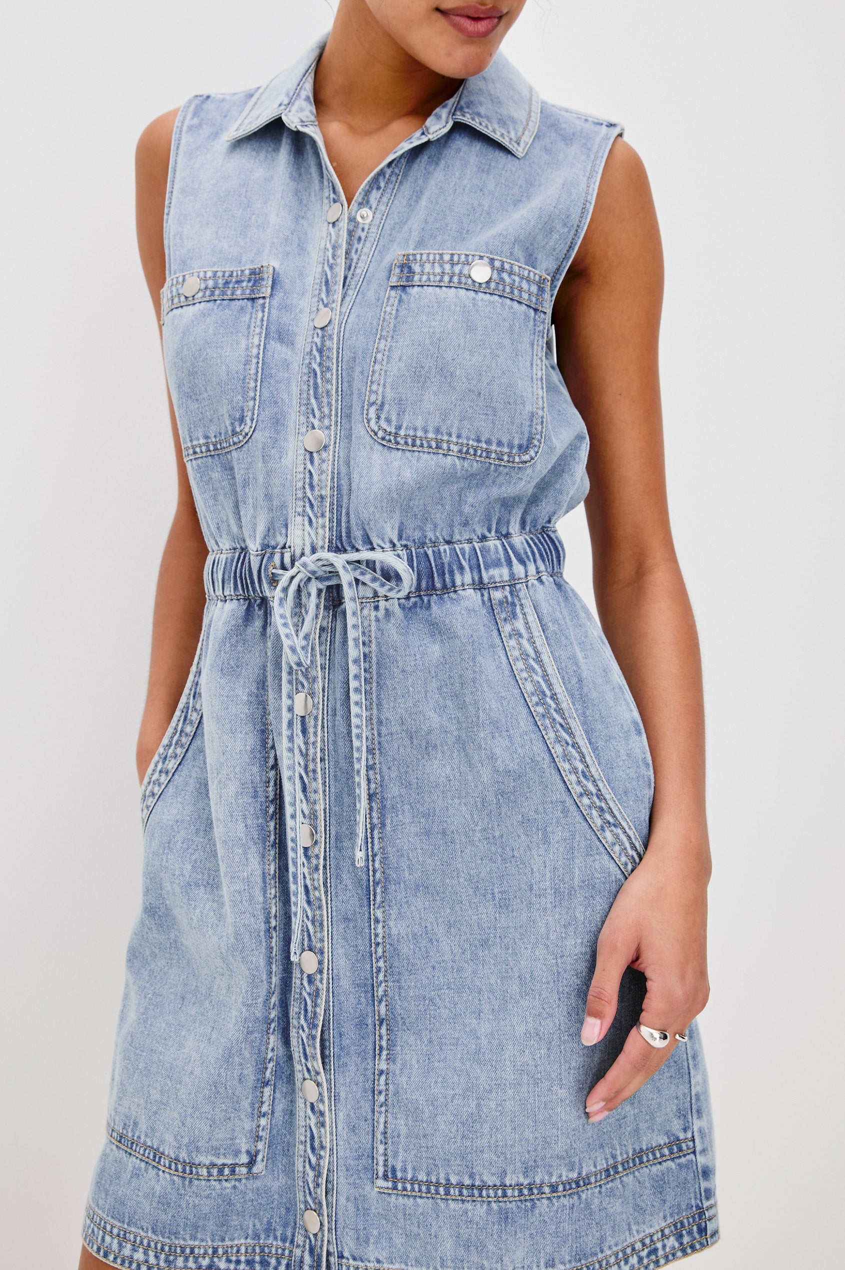 Erica Lightweight Denim Mini Dress with a Exposed Button Front, Encased Drawstring Waistband, Front Patch Pockets, Chest Pockets, Sleeveless and Collared in Faded Indigo Colorway. Front Detail View Shown on Model.